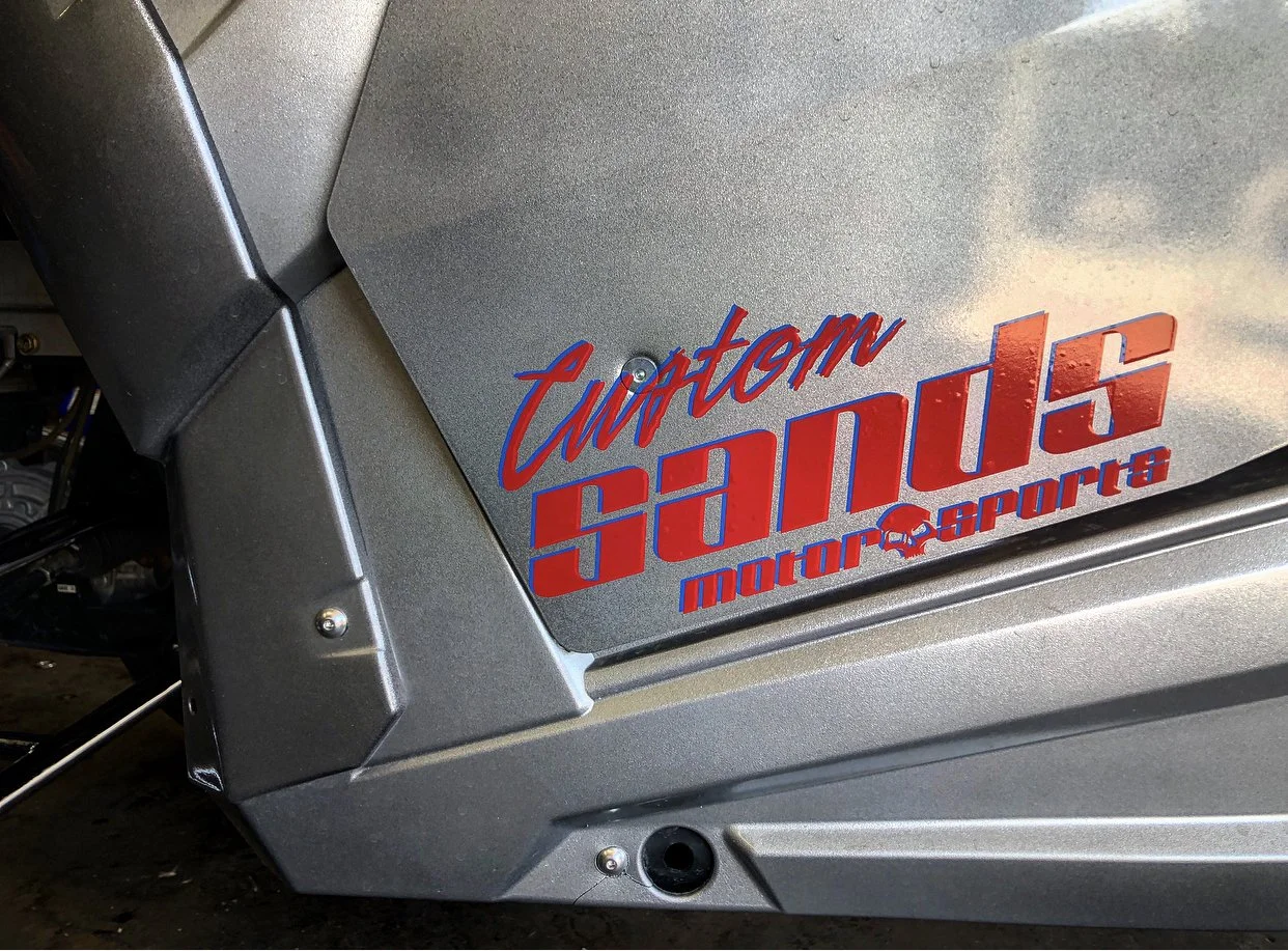 Close-up of a motorcycle side panel with a sticker that reads "Custom Sand Motor Sports" in red and blue lettering.