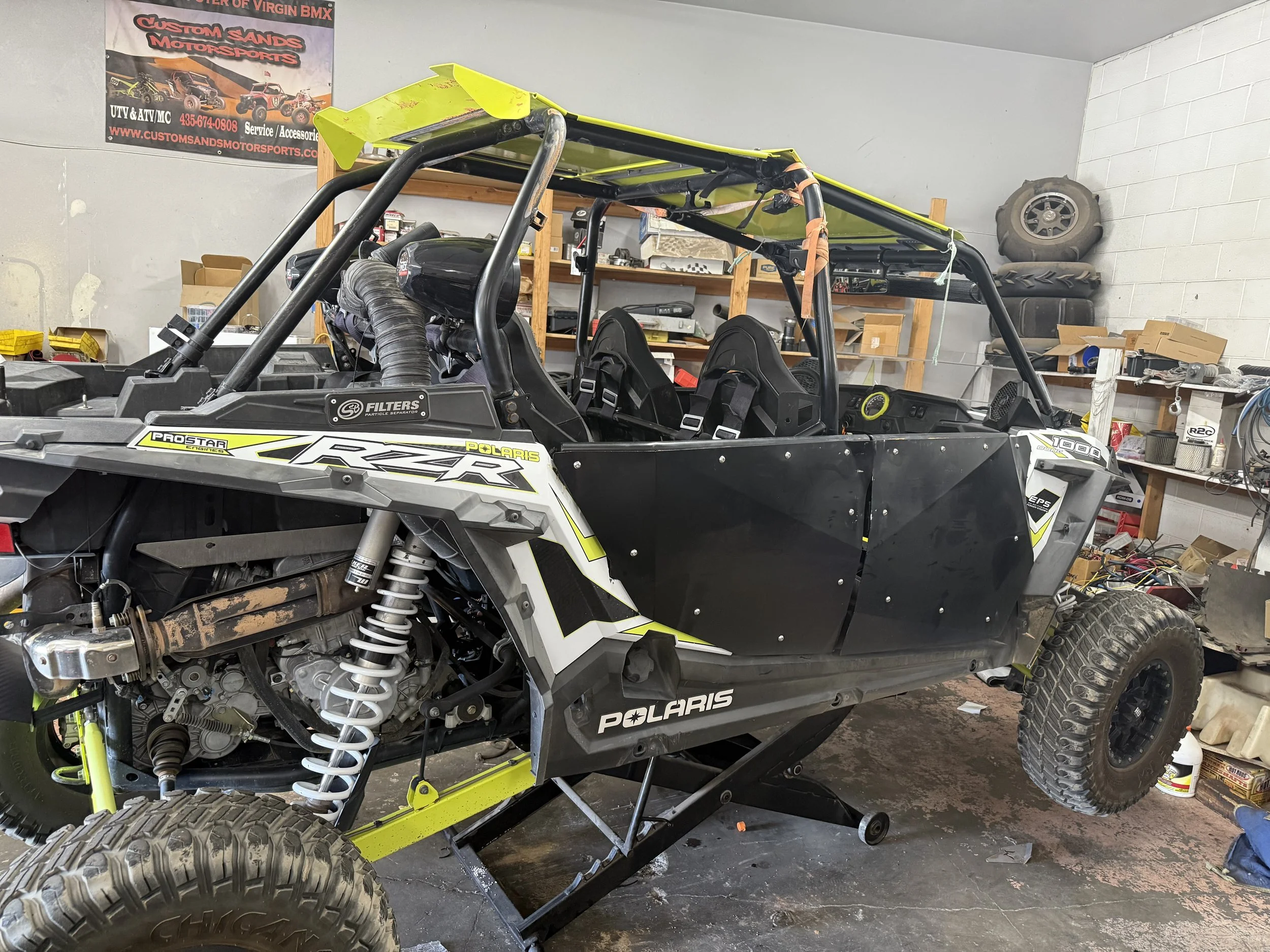 A Polaris RZR side-by-side off-road vehicle inside a garage with parts and tools on shelves and floor.