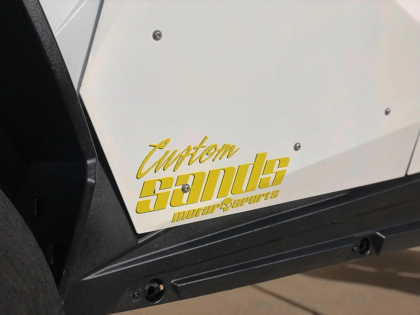 Close-up of a vehicle with a white panel displaying a yellow sticker that reads 'Custom Saints Motor Sports' in stylized lettering.