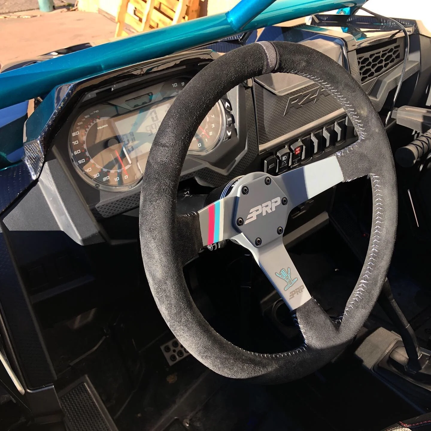 Close-up of a vehicle's dashboard and steering wheel, which is a Momo Pro steering wheel with black suede wrapping and blue, red, and light blue stripes, inside a race or off-road vehicle. The dashboard features a speedometer, tachometer, and various switches.
