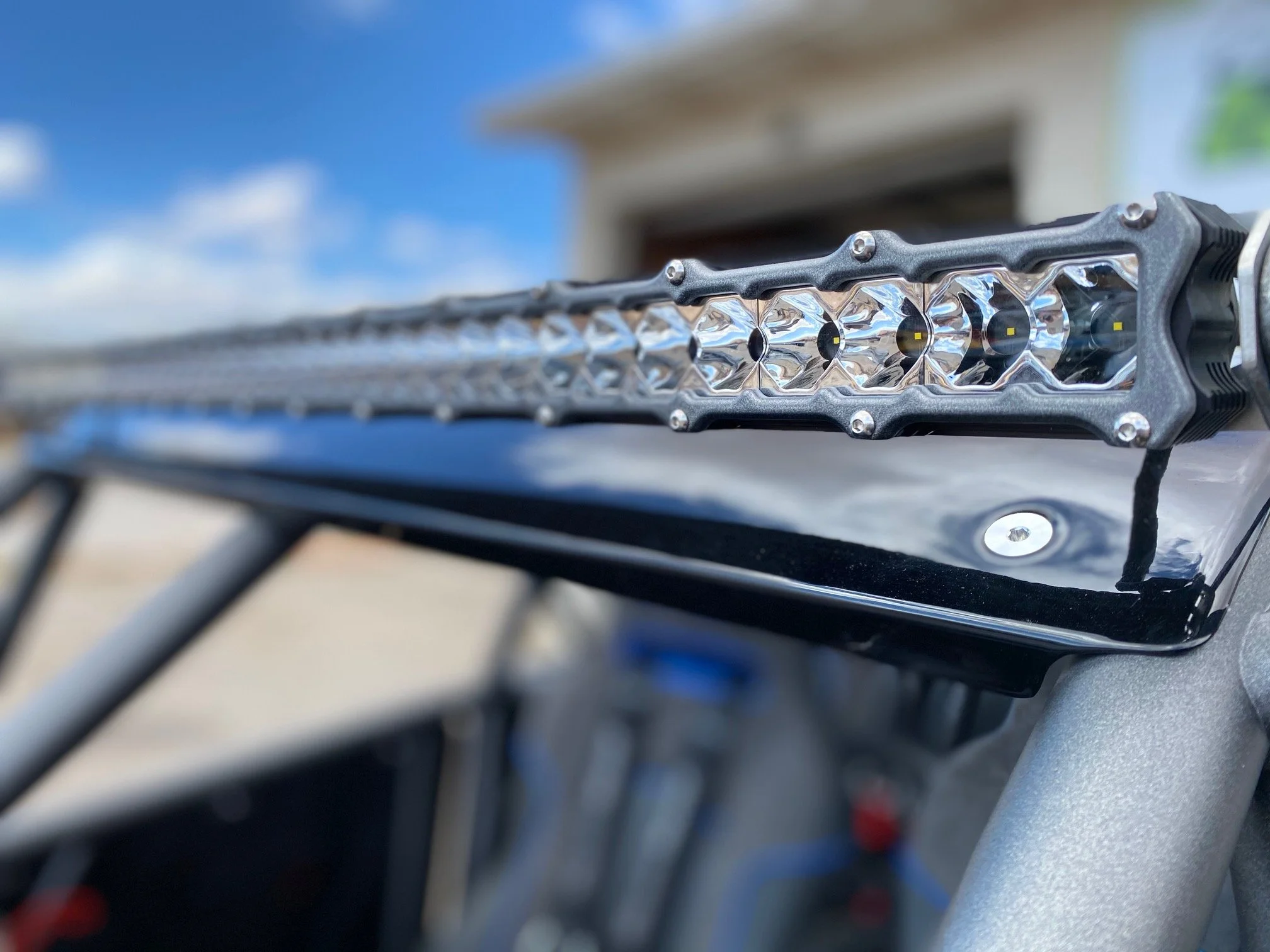 Close-up of an LED light bar mounted on a vehicle with a black surface and a blurred background showing part of a building and blue sky.