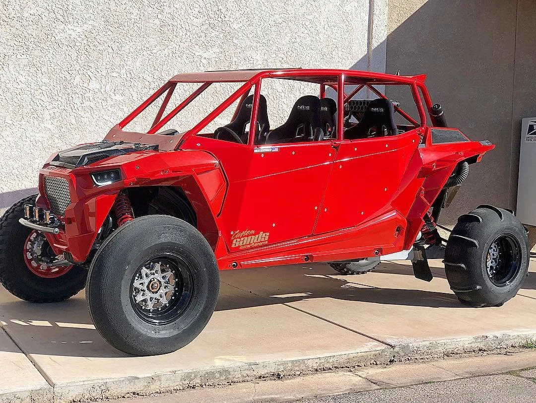 A bright red, open-frame off-road vehicle with four seats and large tires, parked on a concrete surface beside a beige wall.