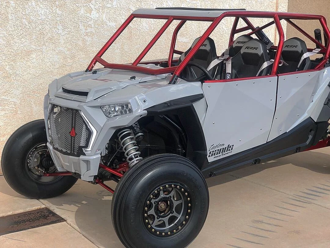 A white and black off-road racing vehicle with a red frame and open seating for four, parked on a sidewalk against a beige stucco wall.