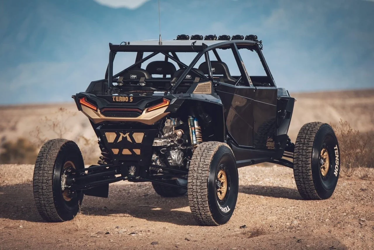 An off-road racing vehicle in a desert landscape, equipped with large tires, a roll cage, and a view of its suspension and engine.