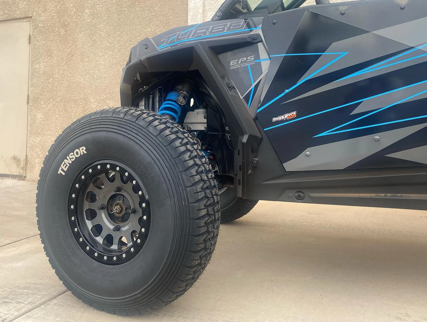 Close-up of the front wheel and suspension of an off-road vehicle, showcasing the tire labeled 'TENSOR' and blue coil shock absorber, with part of the vehicle's black and blue geometric body design visible.