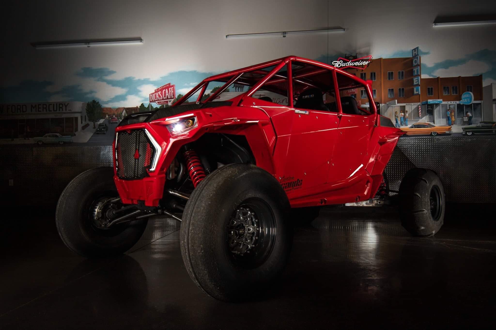 A red off-road racing vehicle with large tires, a roll cage, and exposed suspension, displayed indoors against a mural of a town street scene.