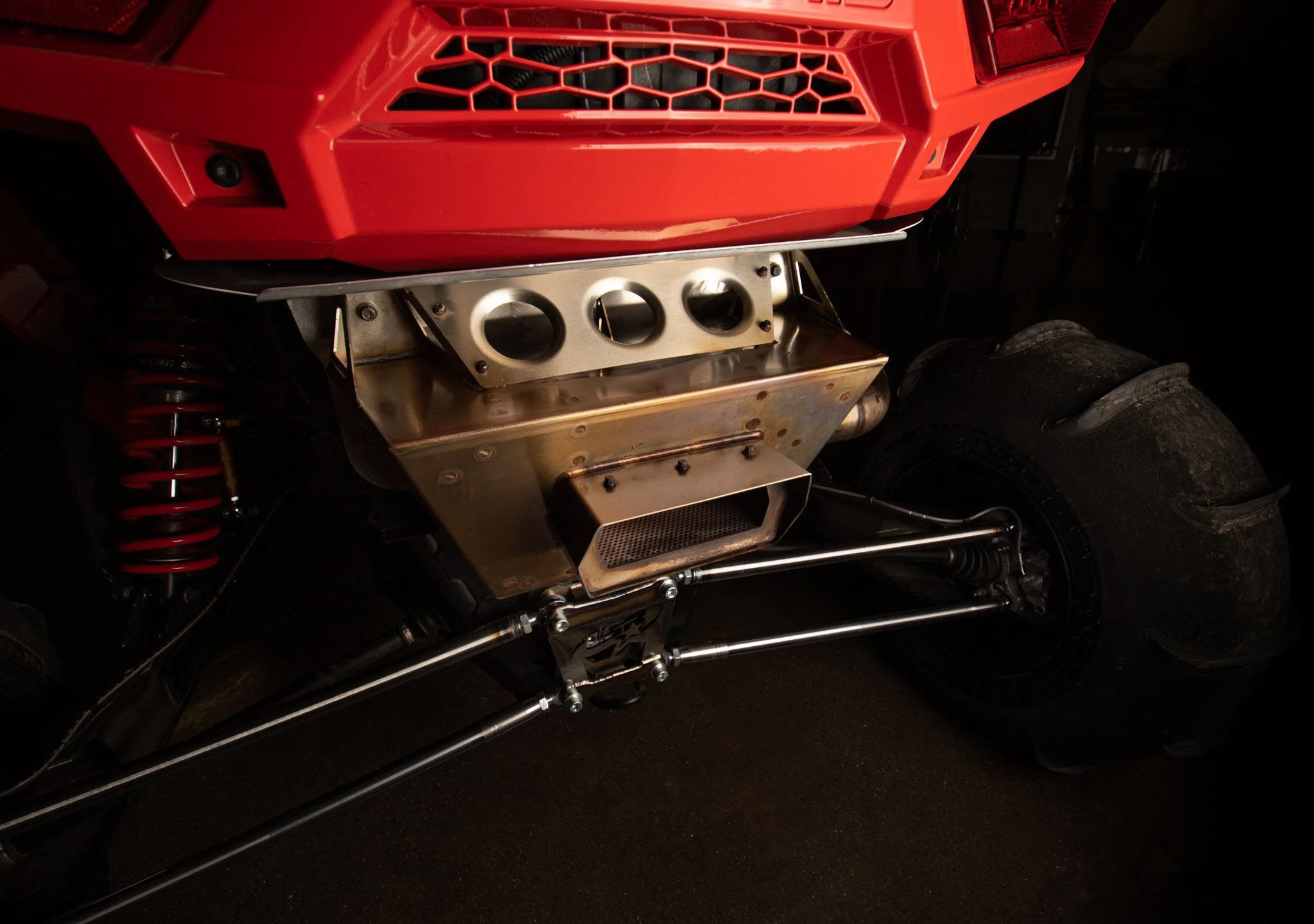 Close-up of a race car's rear, showing a red body, coil suspension, and an exposed, custom-built exhaust system with metallic and mesh components.
