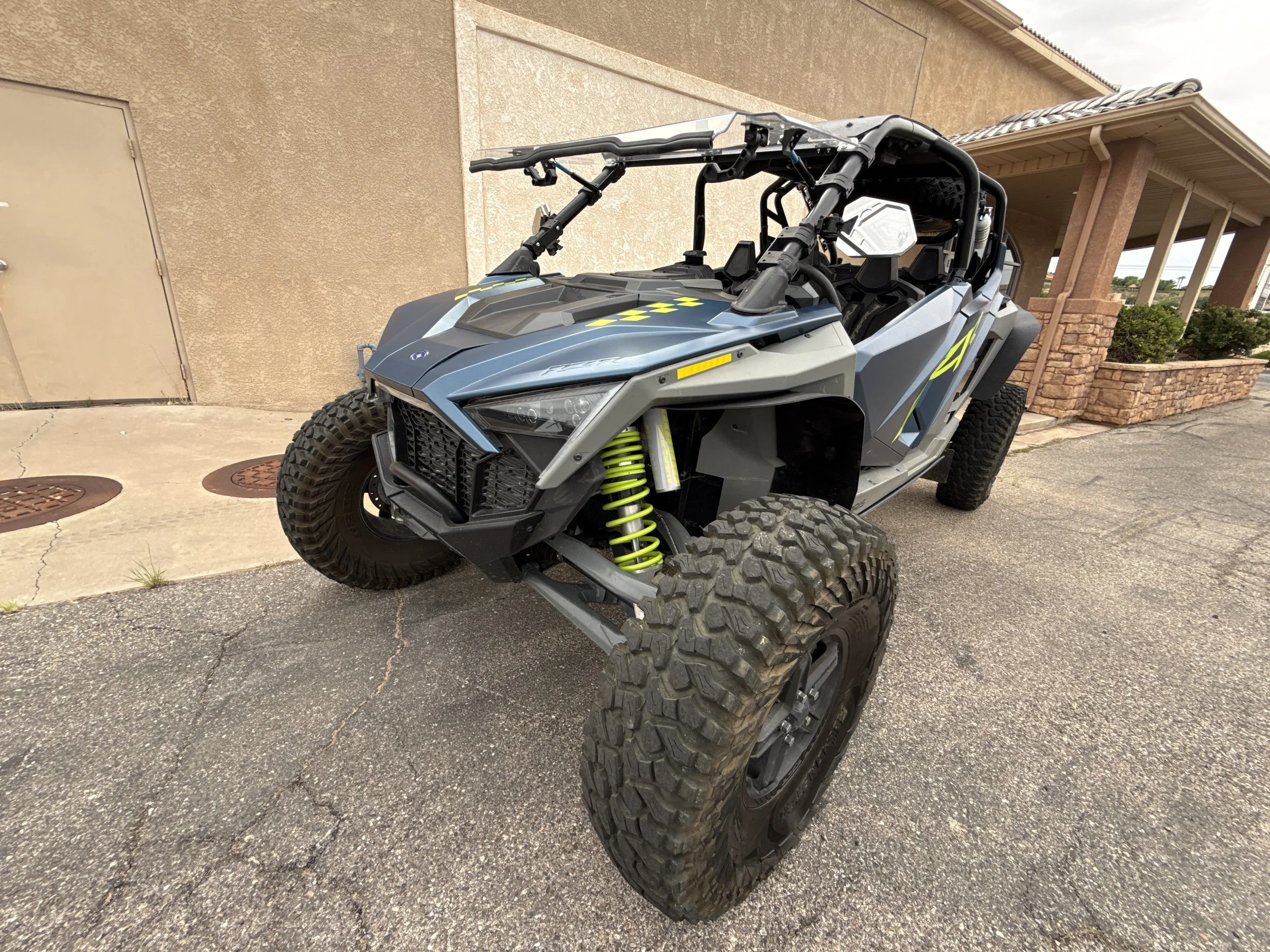 A black off-road dune buggy with large tires, roll cage, and visible suspension system parked on a concrete sidewalk next to a beige wall.