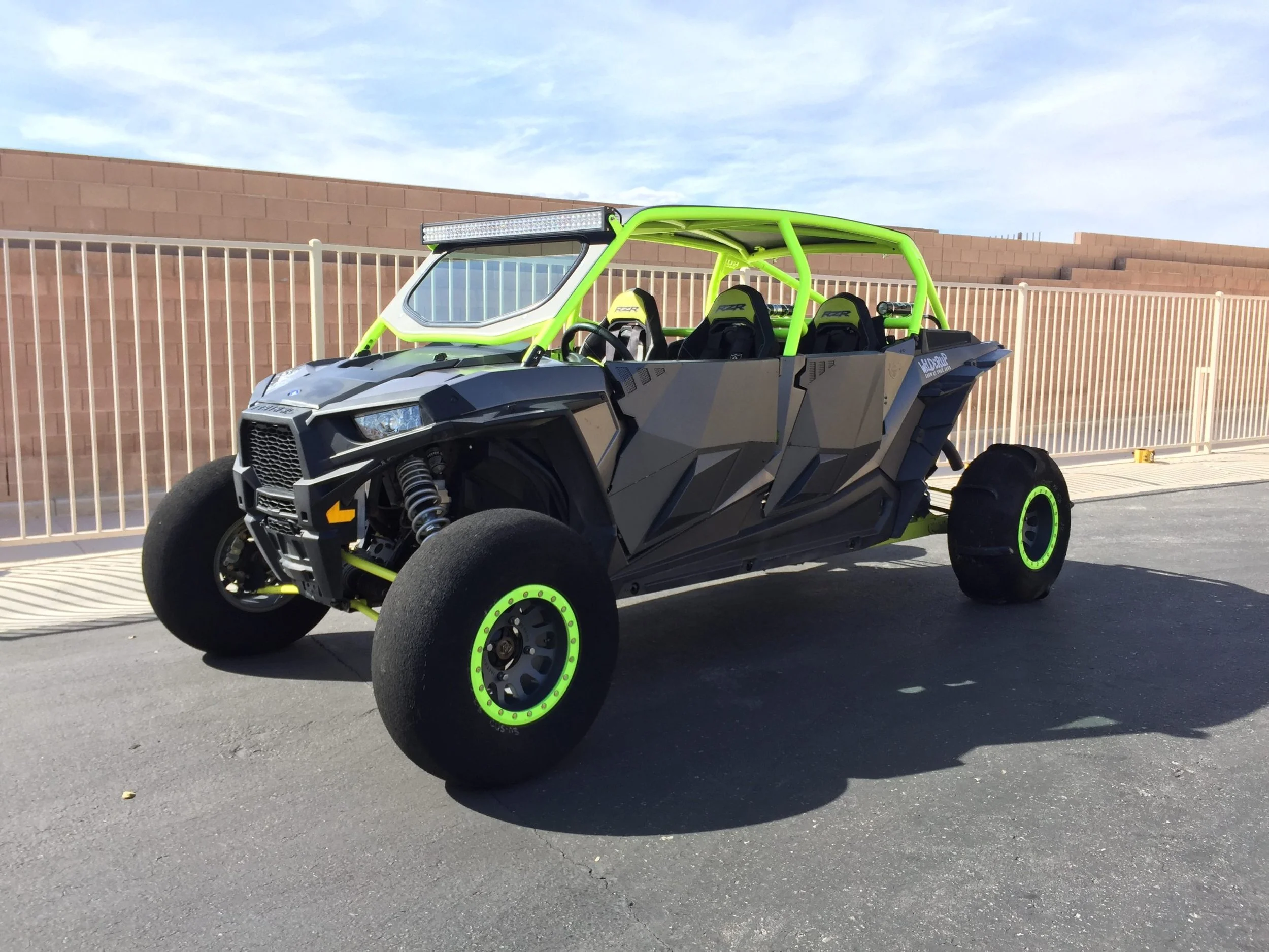 Off-road utility vehicle with a black and neon green color scheme parked on a paved area with a brick wall and fence in the background.