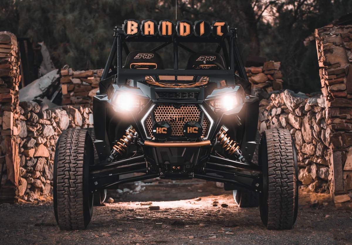 Off-road vehicle with large tires, suspension, and a custom front grille, parked on a dirt path surrounded by stone walls and trees, with a sign reading 'BANDIT' on top.