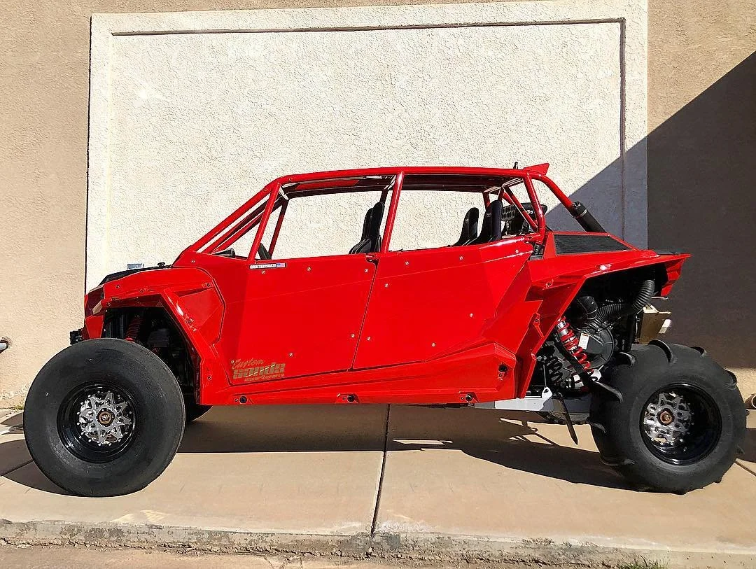 Red off-road dune buggy with large tires, no doors, and black racing seats, parked on concrete in shadowed sunlight.