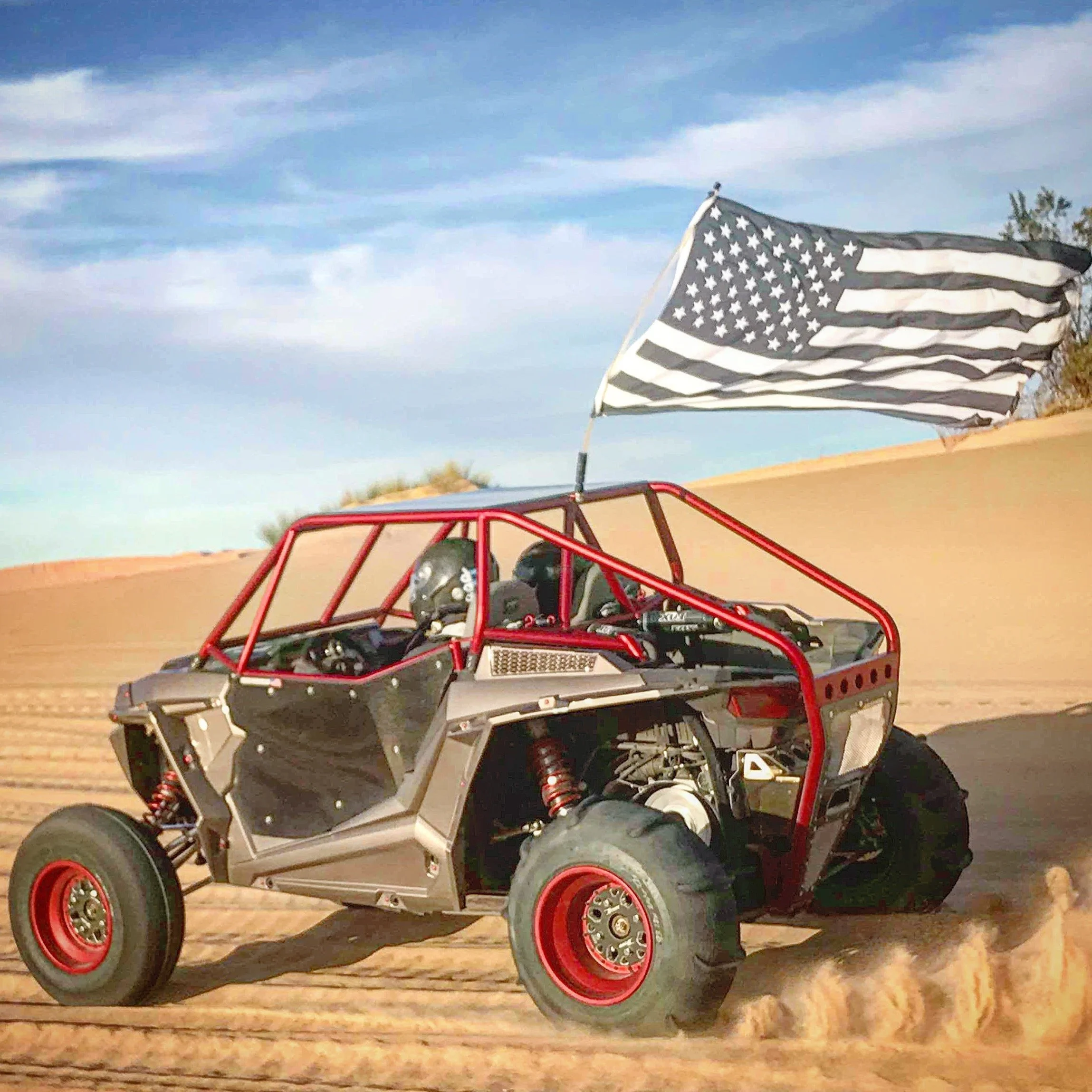 An off-road dune buggy with a red frame and black body, racing across a sandy desert with a black and white American flag attached to the vehicle.