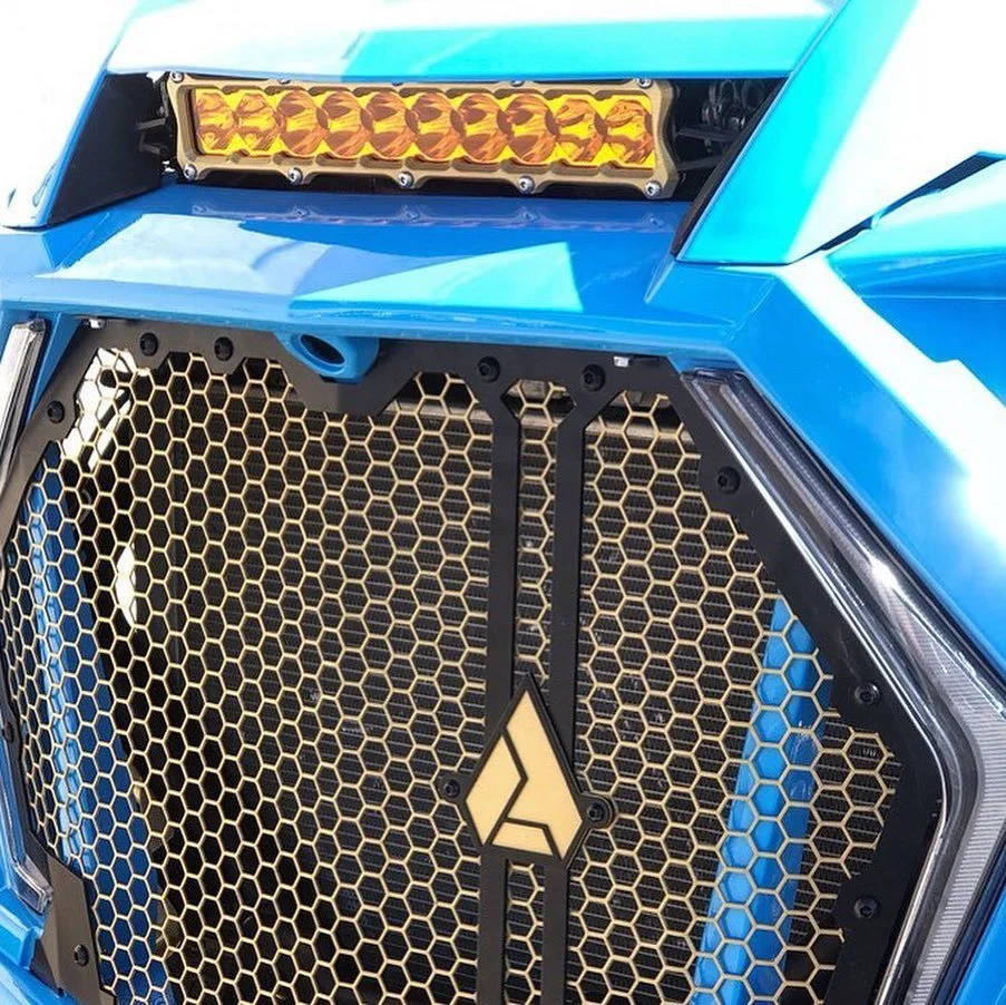Close-up of a blue sports car's front grille with a honeycomb pattern and a black emblem in the center. Above the grille, there is a yellow LED light bar.