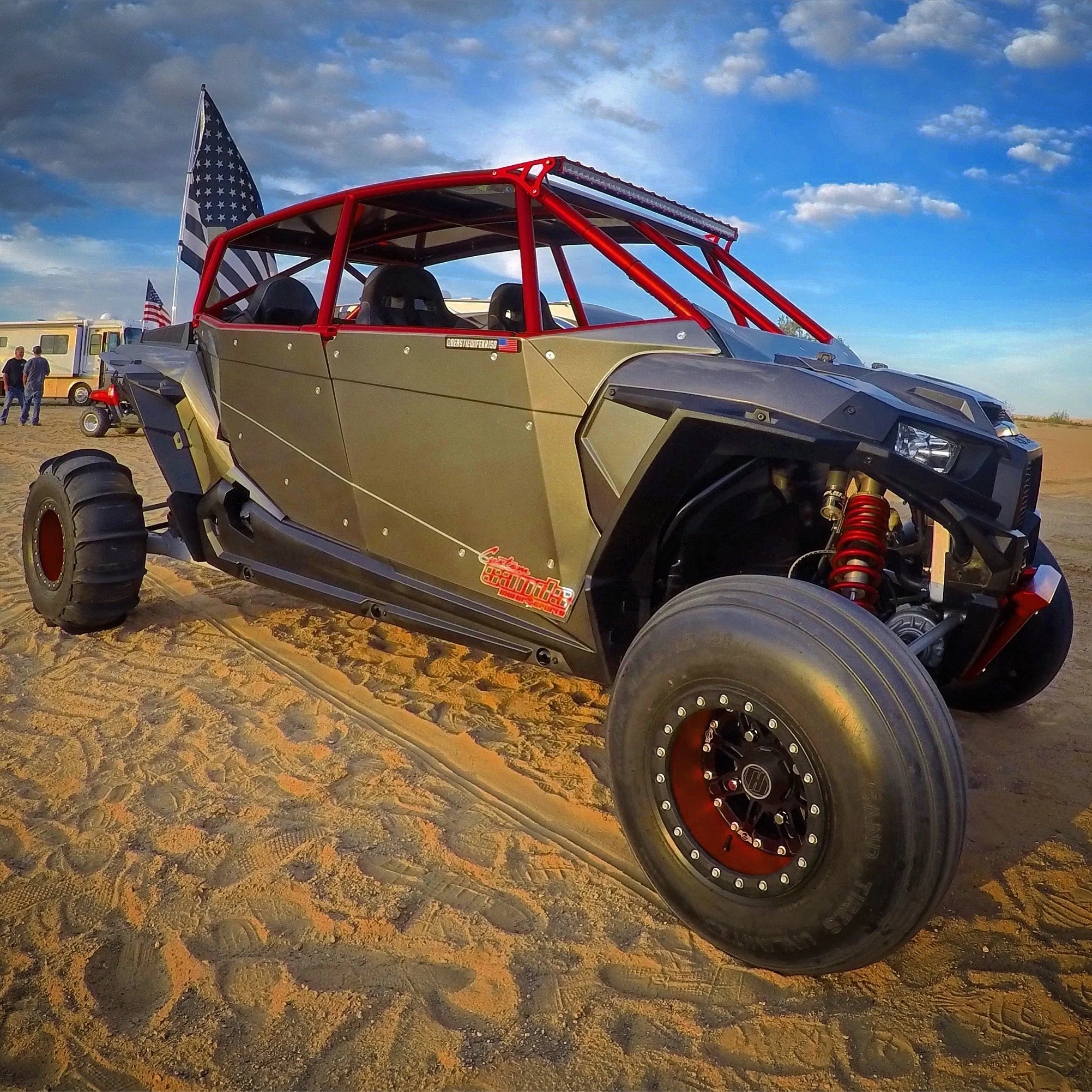 Off-road vehicle with a red roll cage and large tires on sandy terrain, under a partly cloudy sky, with flags in the background.