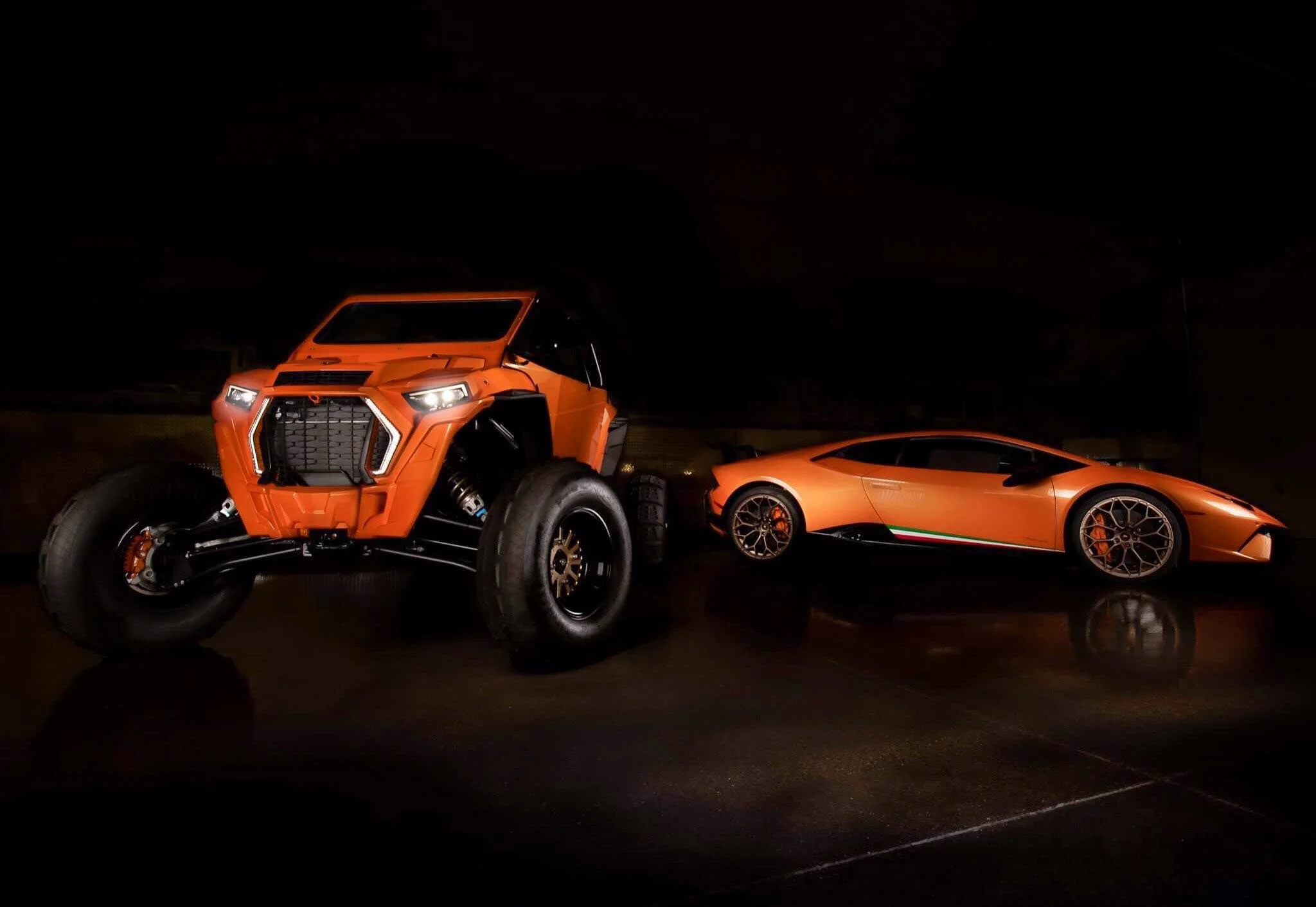 An orange futuristic off-road vehicle with large tires and a sleek sports car with Lamborghini logo parked on a dark surface at night.