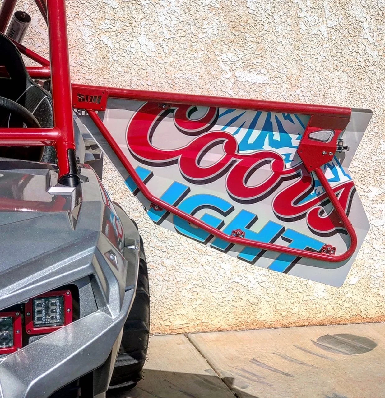 Close-up of a vintage race car with a large, colorful advertising decal on the side, featuring red, blue, and white text. The front of the car shows a silver body and a red frame.