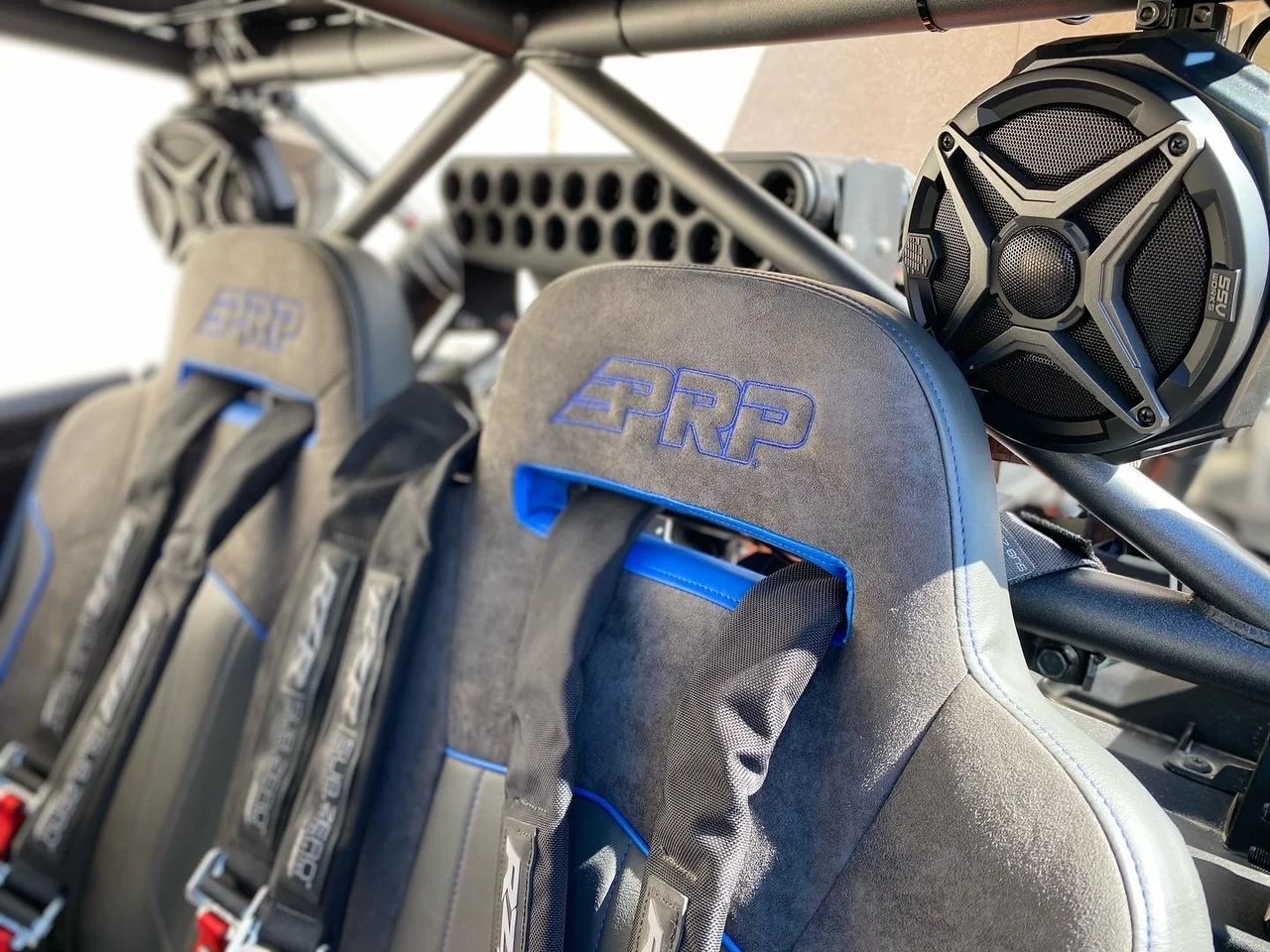 Close-up of racing car seats with 'PRP' logo and harness straps, with a speaker mounted on a metal framework above.