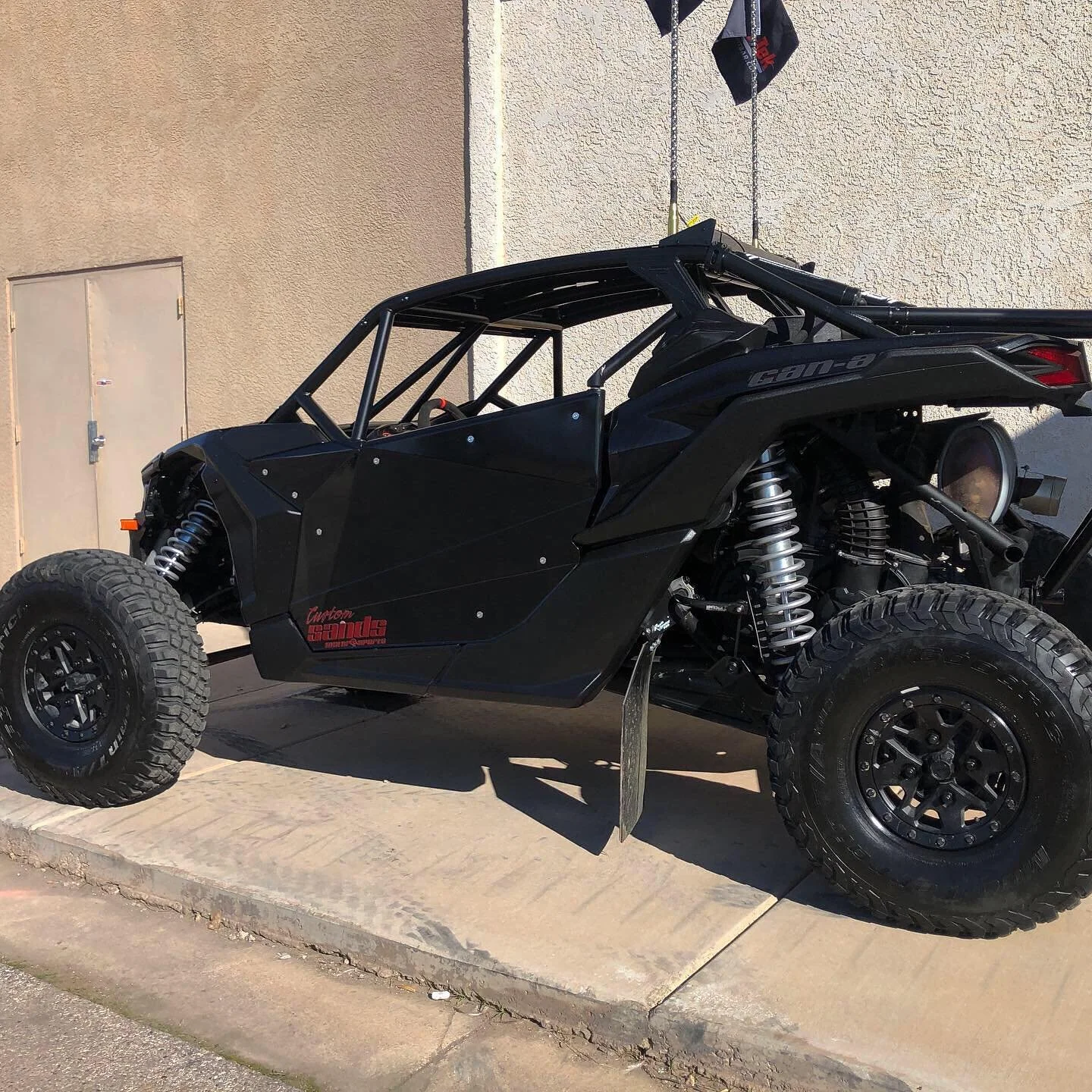 A black off-road dune buggy with large tires, roll cage, and visible suspension system parked on a concrete sidewalk next to a beige wall.
