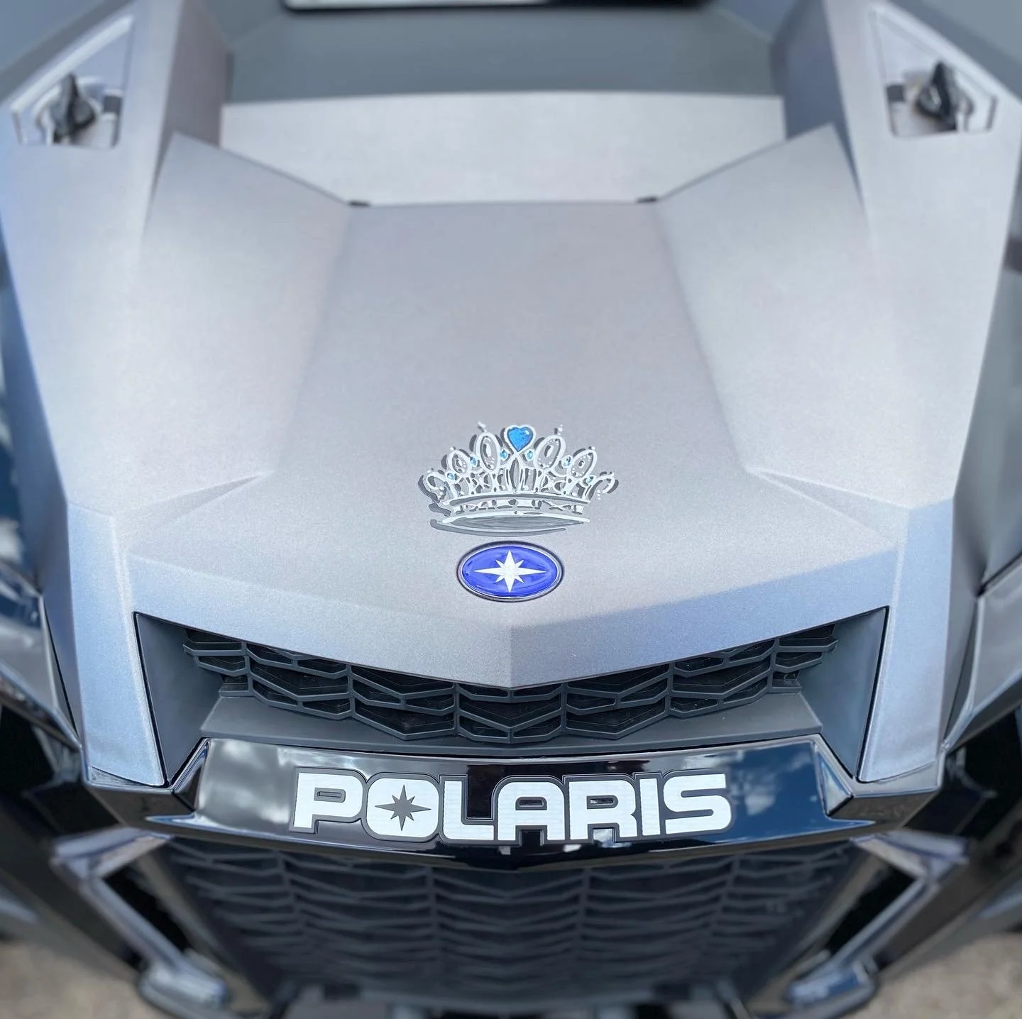 Close-up of the front of a silver Polaris vehicle, featuring a crown decal with a blue heart, the Polaris logo, and a custom license plate reading 'POLARIS'.