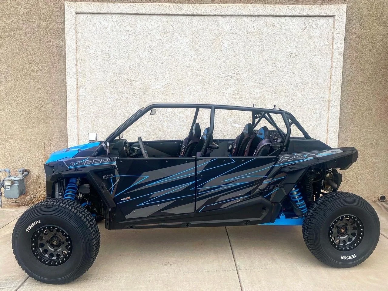 A black and blue off-road dune buggy with four seats, large tires, and visible suspension system is parked on a concrete driveway next to a beige wall.