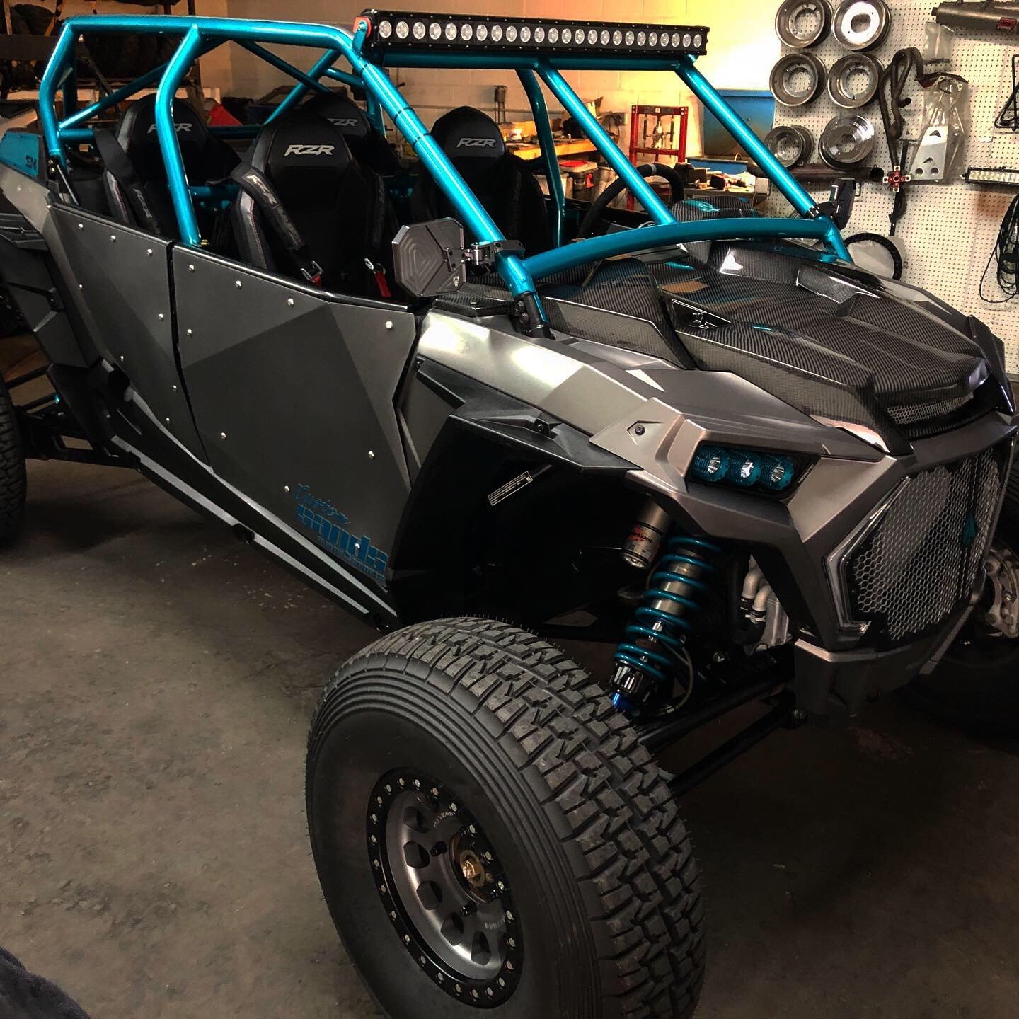A rugged, open-frame off-road vehicle with black and gray body panels, large off-road tires, blue suspension springs, and two racing seats inside, parked in a garage with tools on a pegboard in the background.