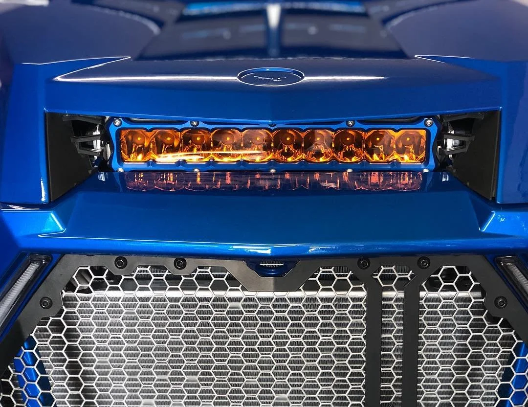 Close-up of the front of a blue sports car, featuring an orange LED light bar and a honeycomb grille.