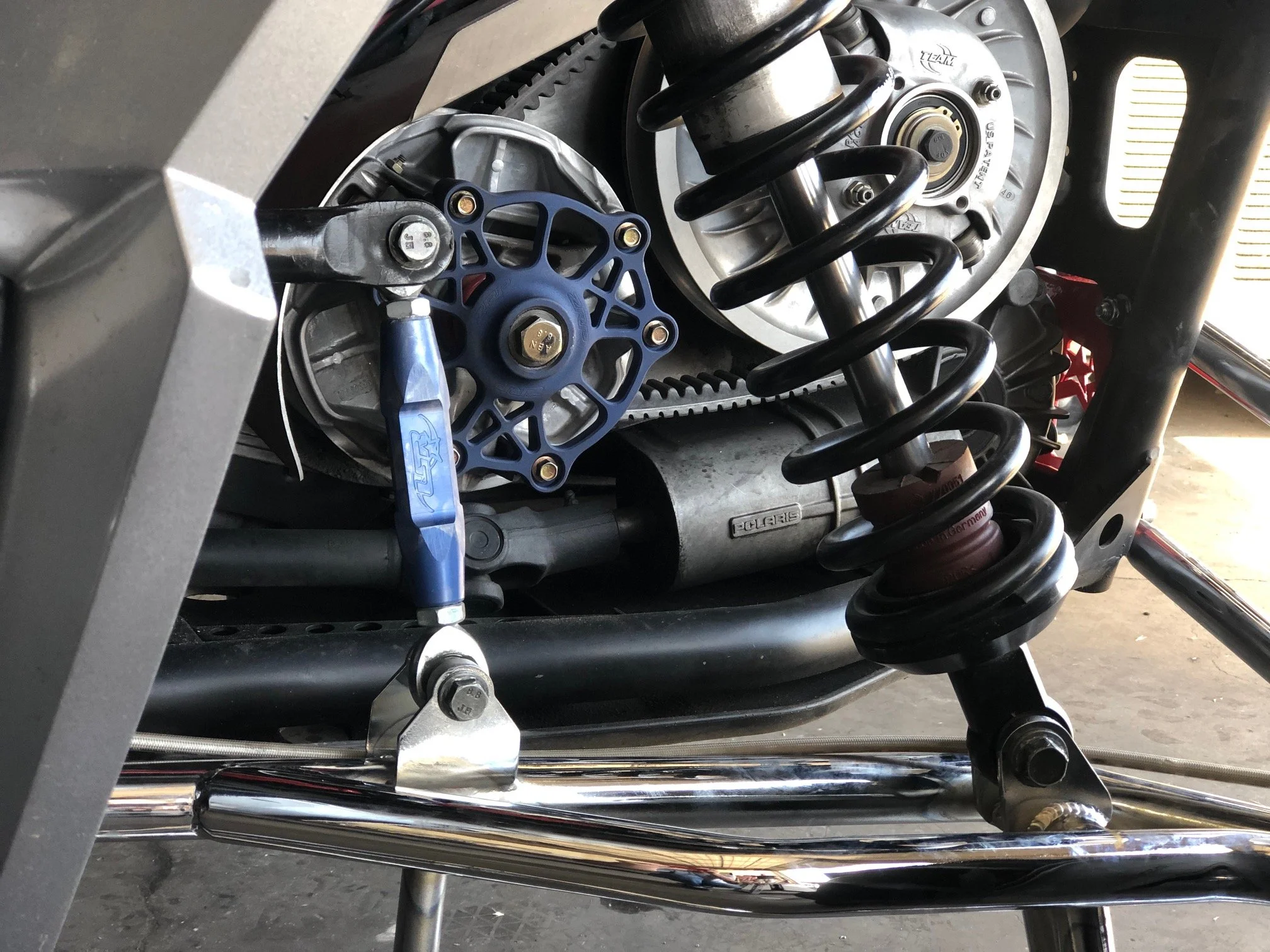 Close-up of a motorcycle suspension and engine parts, including a coil spring, pulley, and belt, with metal components and bolts.