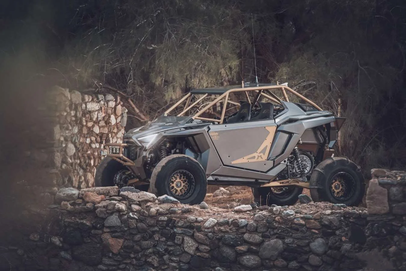 A rugged off-road electric vehicle with a metallic exterior, exposed frame, and four large tires, parked among rocks and stone structures at night.