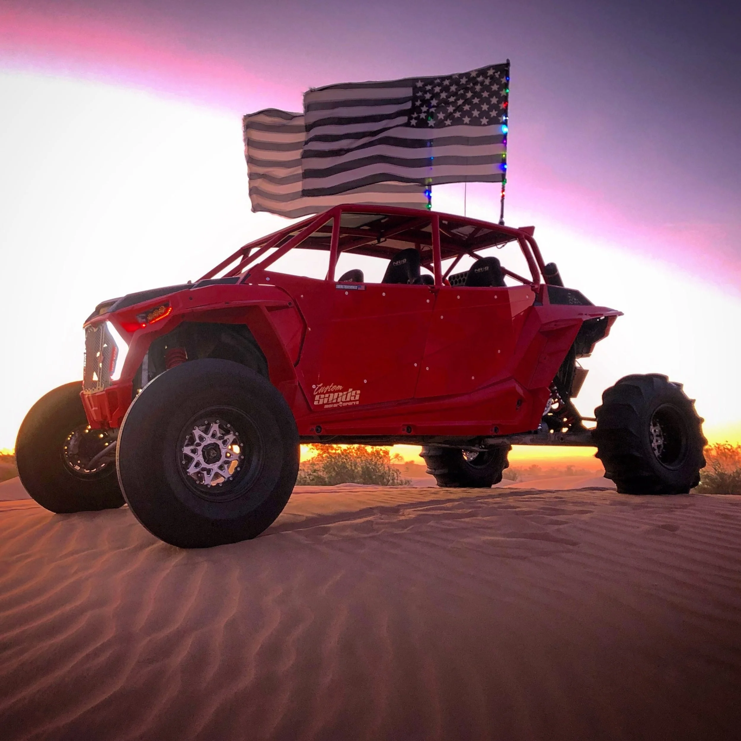 A red off-road vehicle with large tires parked on sandy terrain during sunset with two American flags mounted on top, one of which is illuminated with colorful lights.