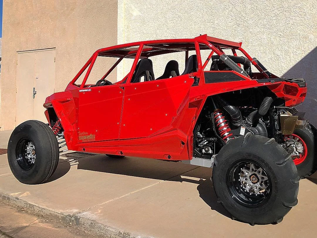 A partially assembled off-road race car with a red body and four large tires, parked on a concrete surface with a beige wall in the background.