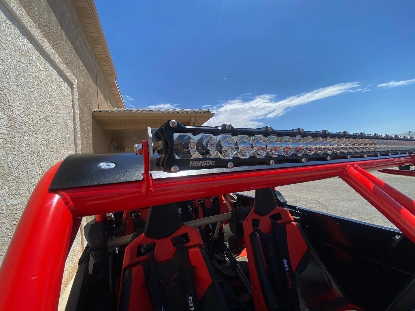Close-up of a red off-road vehicle with a black roof and roll cage, featuring a large LED light bar mounted on top, set against a bright blue sky with some clouds.