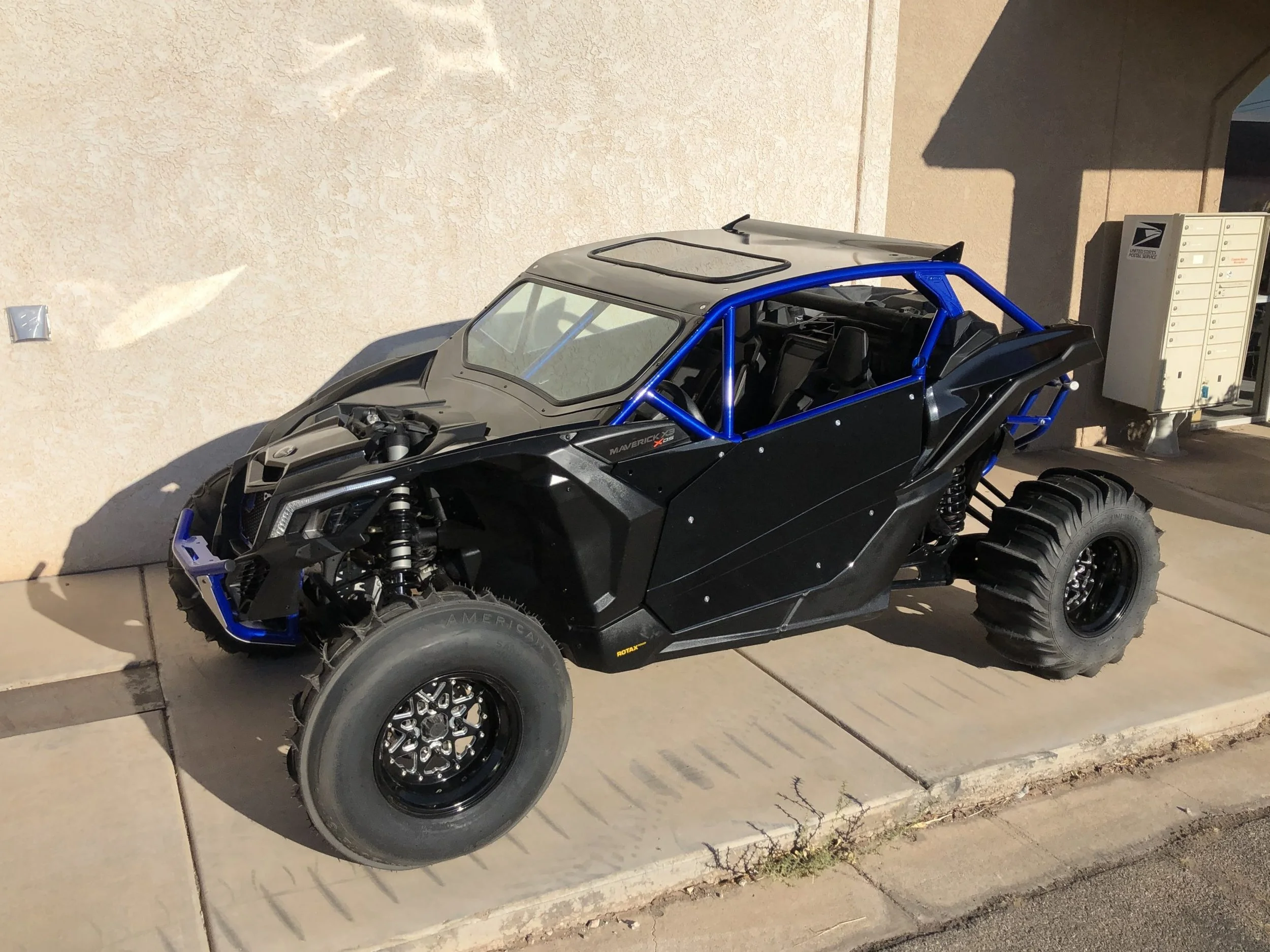 Off-road buggy parked on a sidewalk next to a beige wall with visible electrical boxes. The buggy has large tires, a black body with blue roll cage, and an open interior.