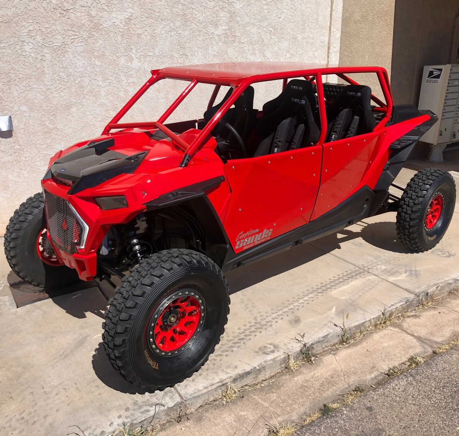 A red off-road dune buggy with four seats, black racing seats, large off-road tires, and safety roll cage, parked on a concrete sidewalk against a beige stucco wall.