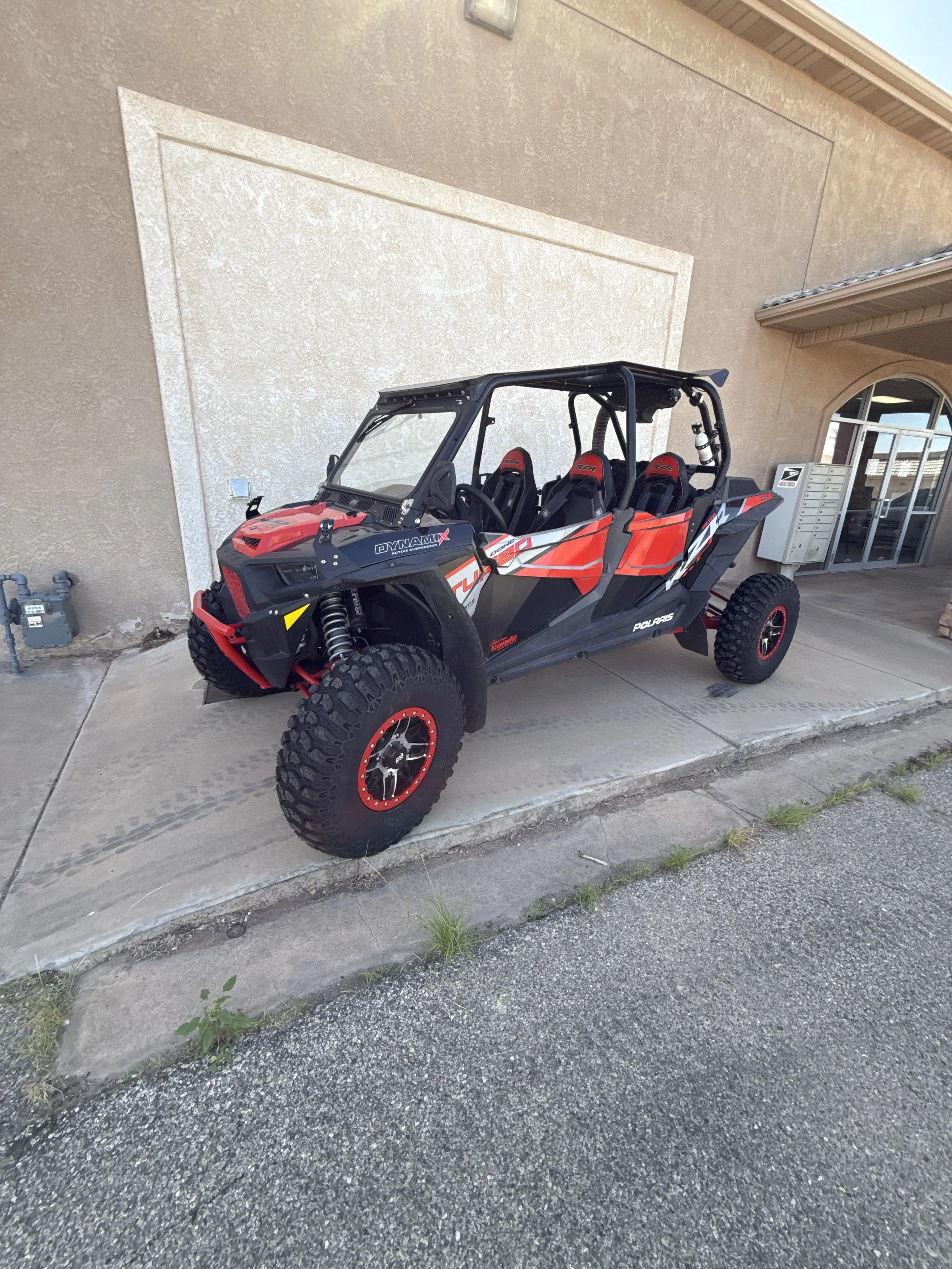 Close-up of a black off-road vehicle with a prominent grille and Suspension, parked in front of a business sign that reads Custom Sans Motor Sports, specializing in custom off-road vehicles.