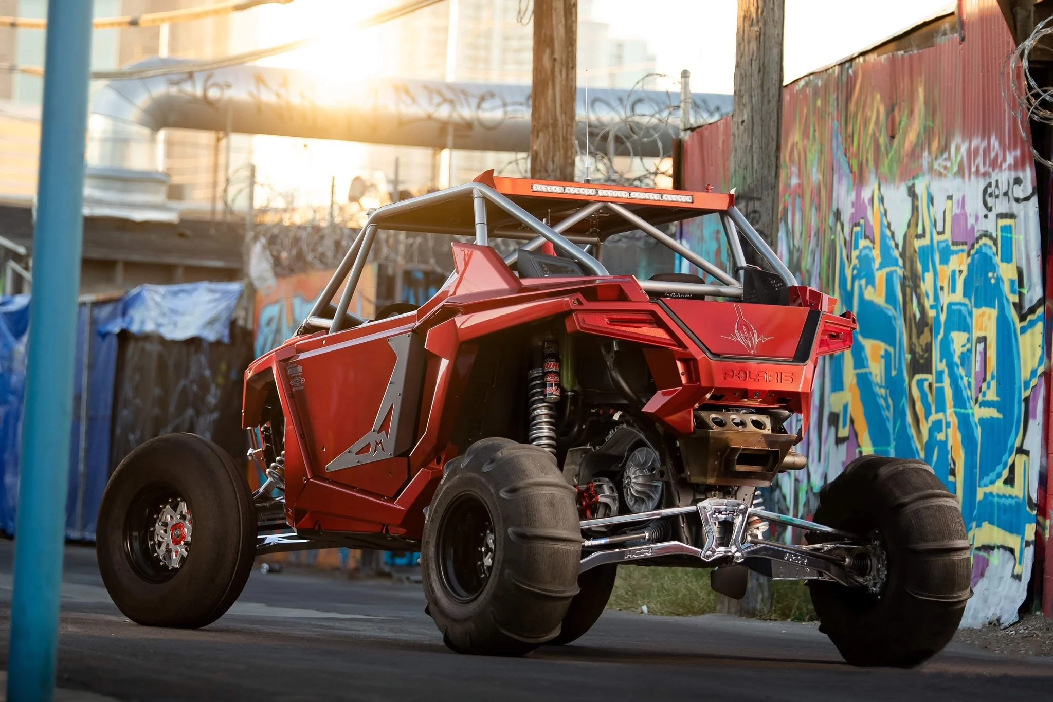 A red Polaris off-road vehicle with exposed engine and large tires, parked outdoors near a graffiti-covered wall during sunset.