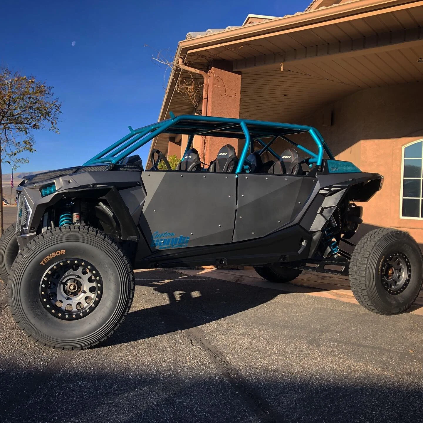 A black off-road dune buggy with blue roll cage parked outside a tan building with a window, under clear blue sky with the moon visible.
