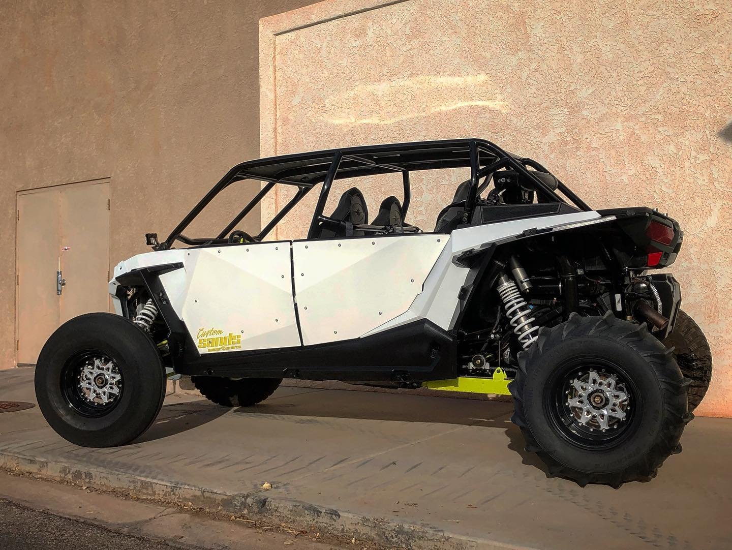 A custom off-road dune buggy with large tires, exposed suspension, and a roll cage, parked on a sidewalk at night.