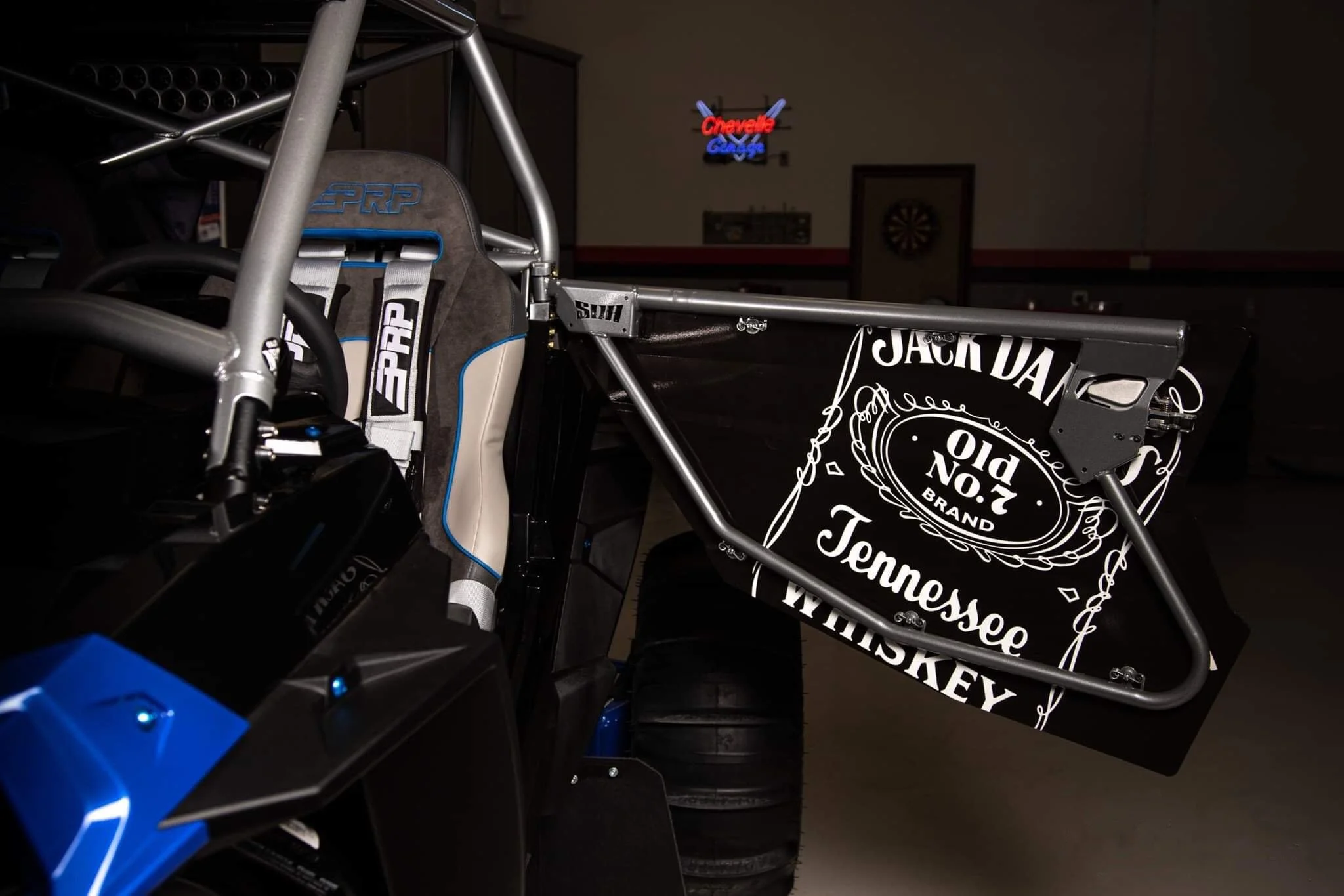 Close-up view of a race car cockpit showing a racing seat with PRP branding, a metal roll cage, and a black rear wing with Jack Daniel's Tennessee Whiskey branding in a dimly lit indoor setting.