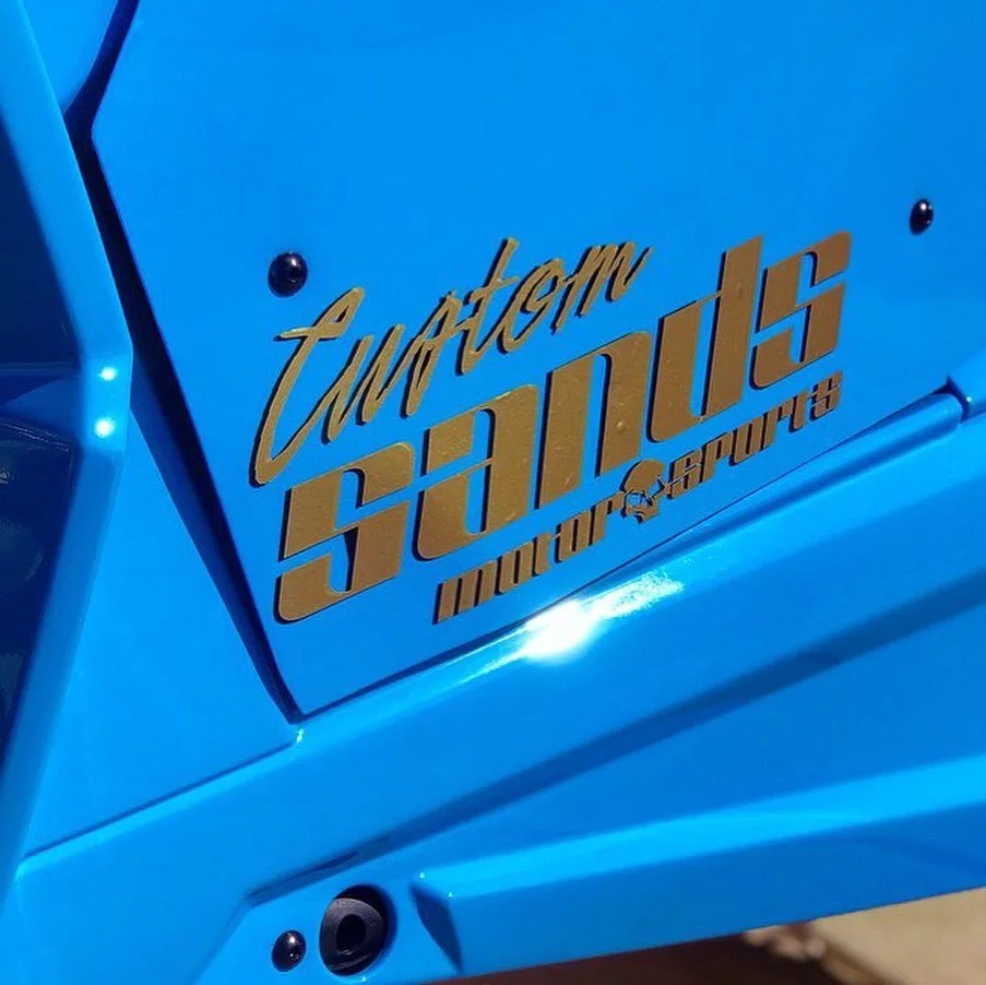 Close-up of a blue vehicle with a decal that reads 'Custom SouLutions MotorSports' in gold lettering.