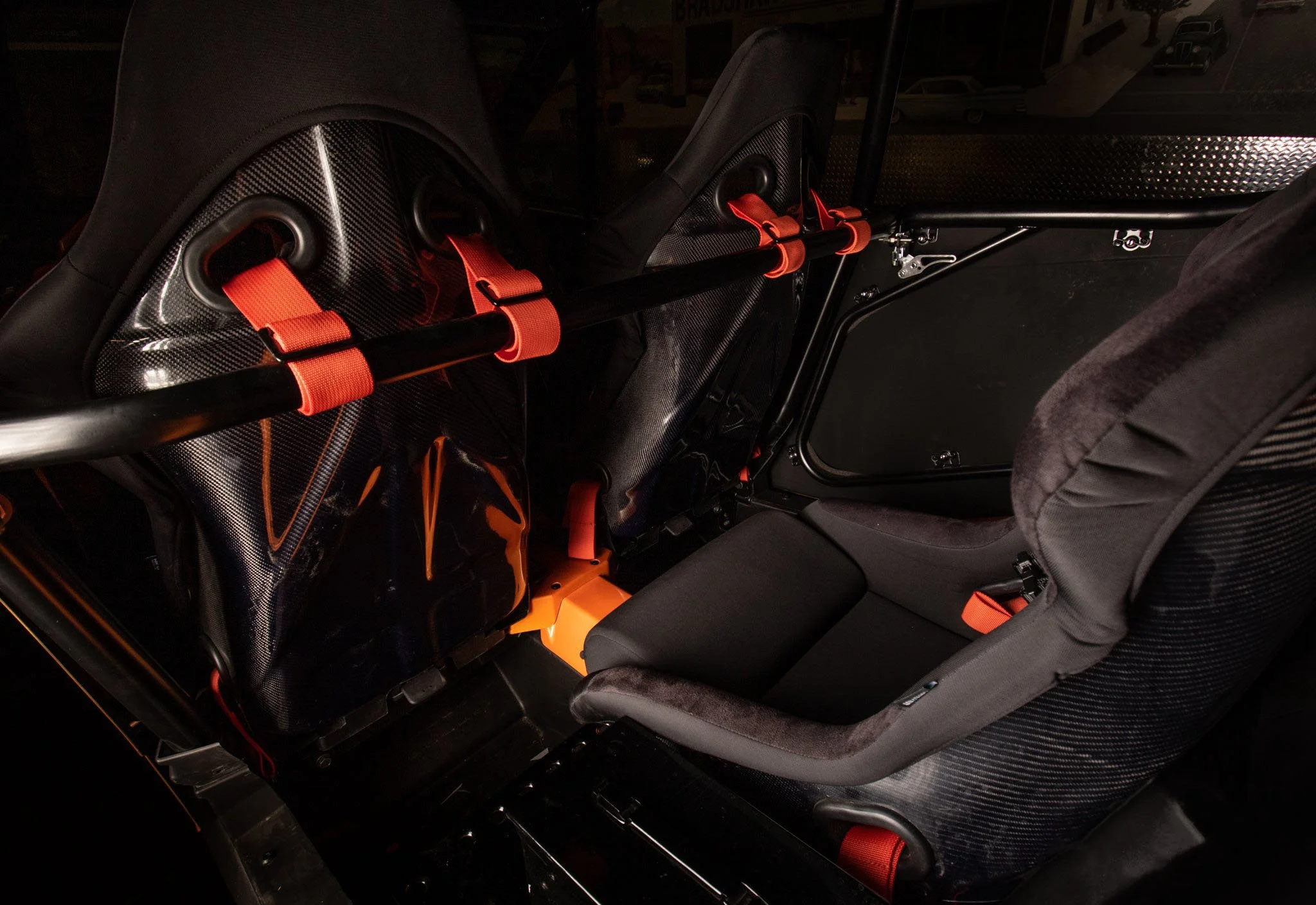Interior of a race car with black and orange safety harnesses and seats, black carbon fiber panels, and a steel roll cage.
