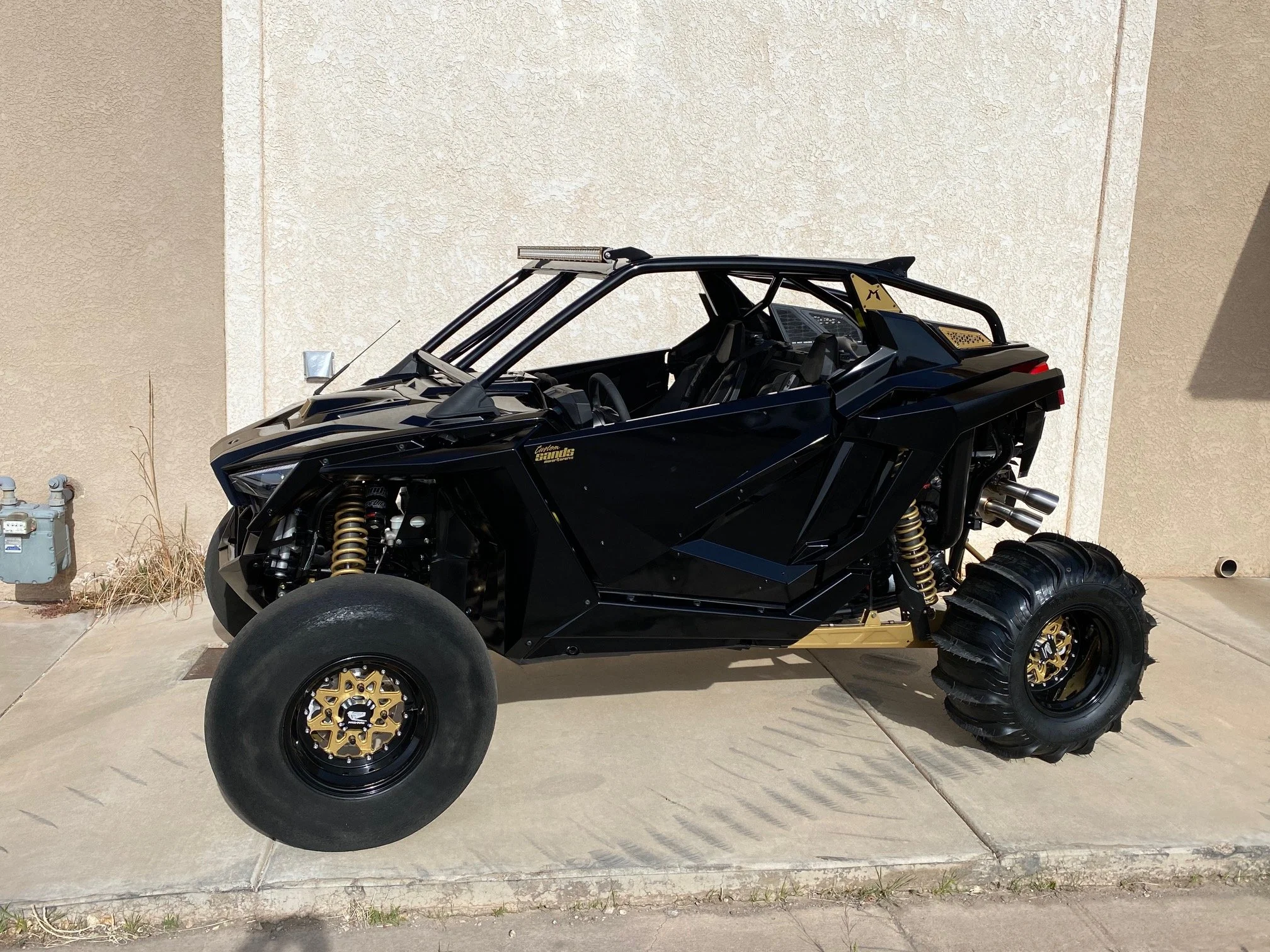 A custom black off-road vehicle with gold accents, large tires, and exposed suspension, parked on a concrete driveway against a beige wall.