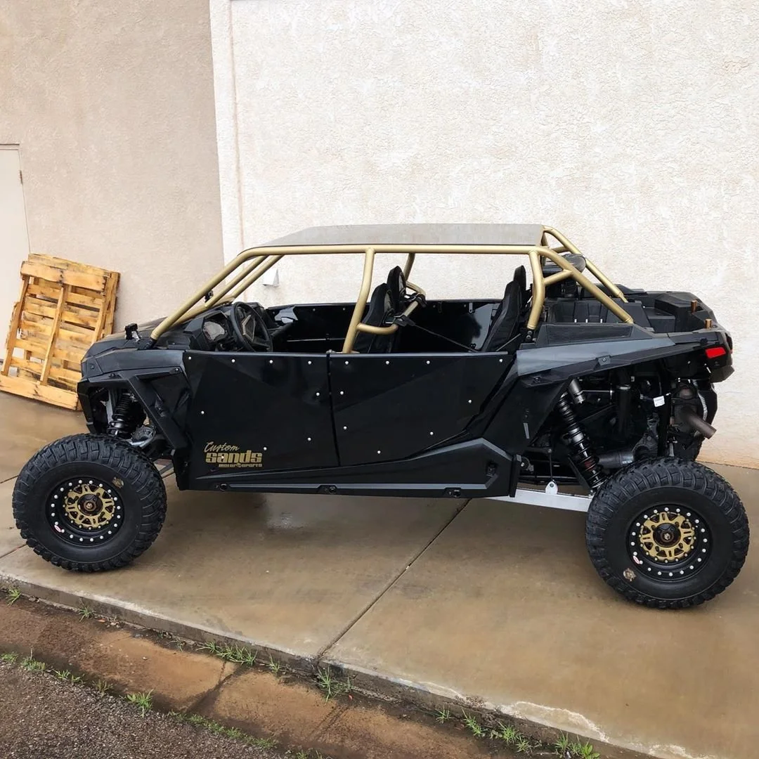 A black off-road vehicle with a gold roll cage and large tires is parked on a concrete surface against a beige wall.