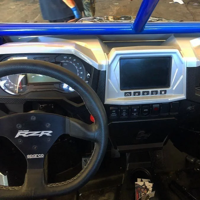 Inside the cockpit of a Polaris RZR off-road vehicle, showing the steering wheel, dashboard with speedometer, and a central display screen.