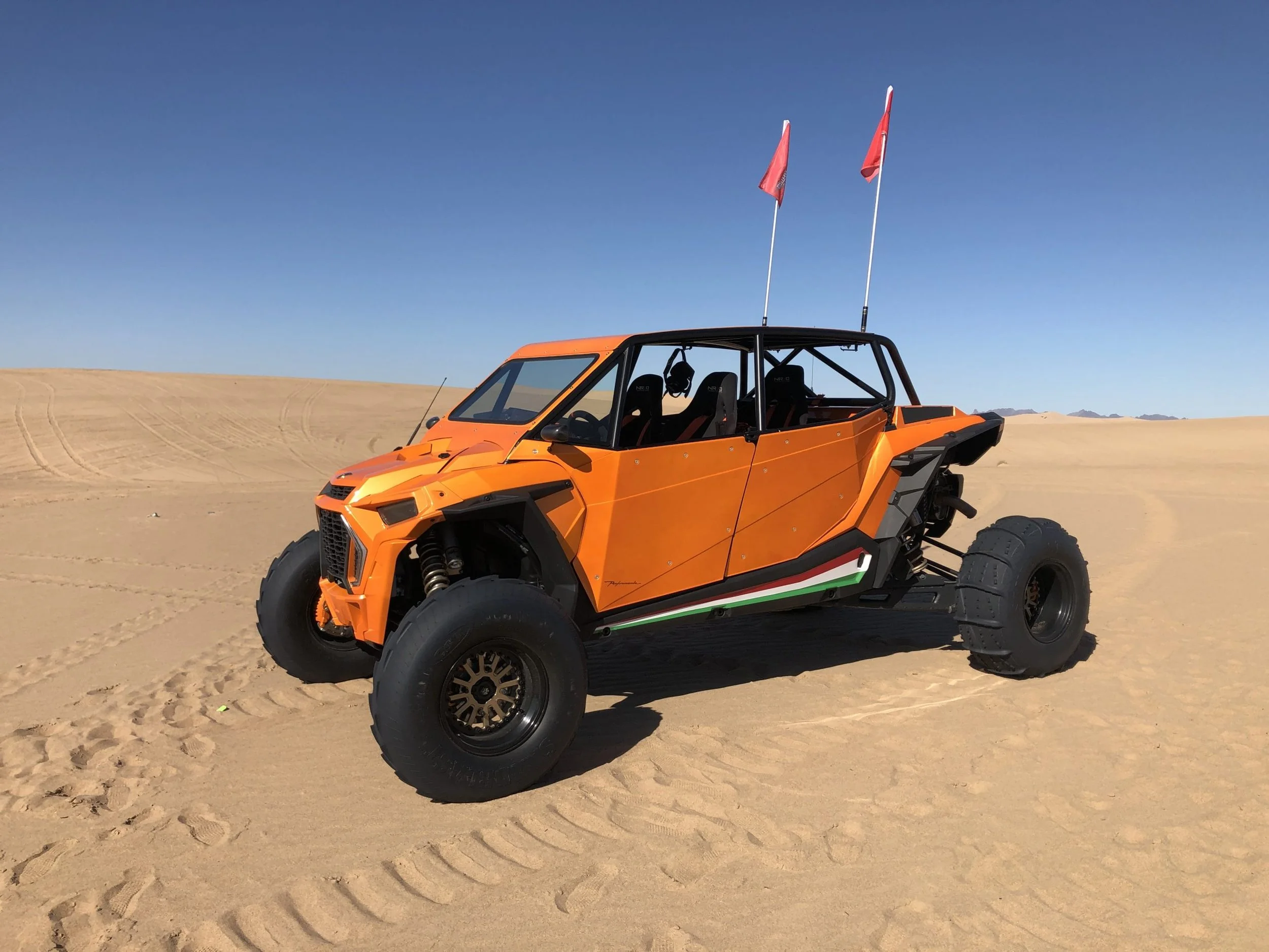 An orange off-road vehicle with four large tires and a roll cage, parked on sandy terrain under a clear blue sky, with two orange flags mounted on top.
