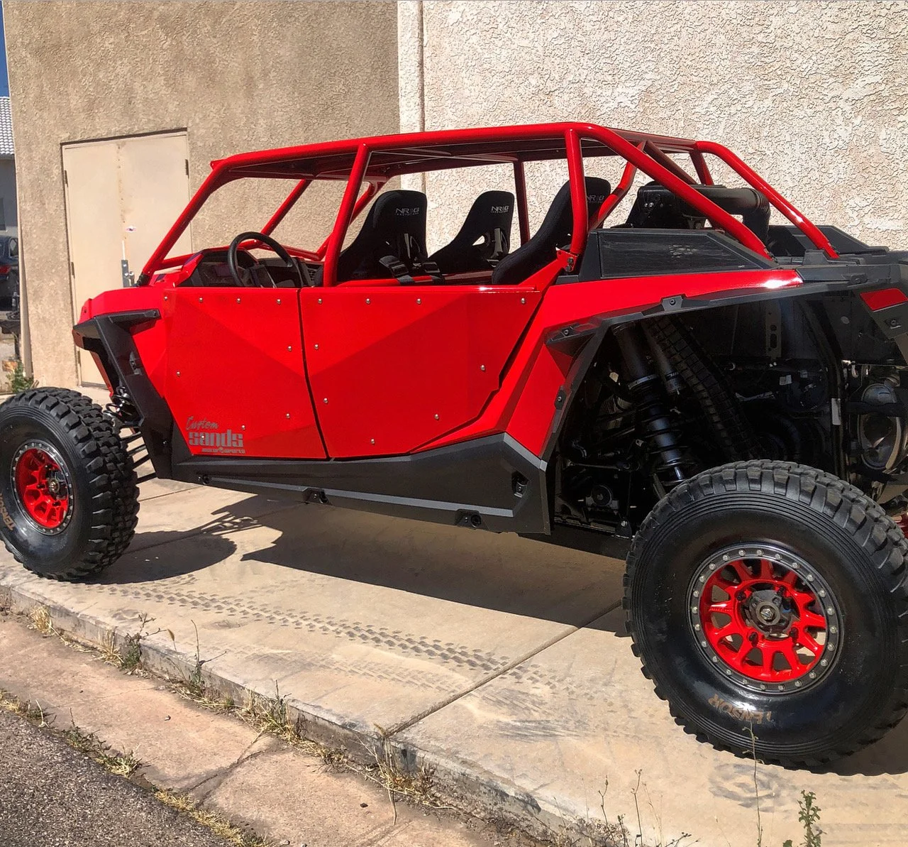 A red off-road dune buggy with black tires and a roll cage, parked on a concrete sidewalk next to a beige wall.