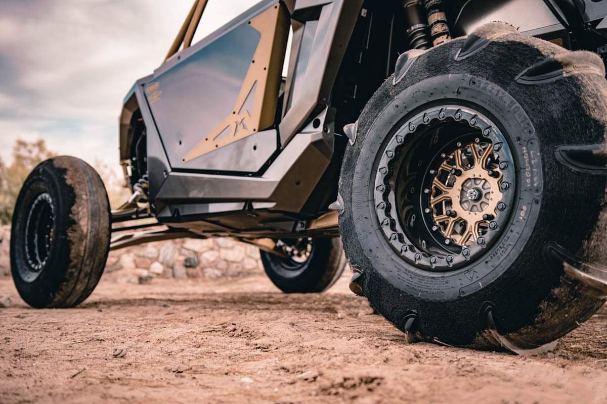 Close-up of rugged off-road vehicle tires on dirt ground, with one tire partially deflated and mud on the tires, and part of the vehicle's body visible.