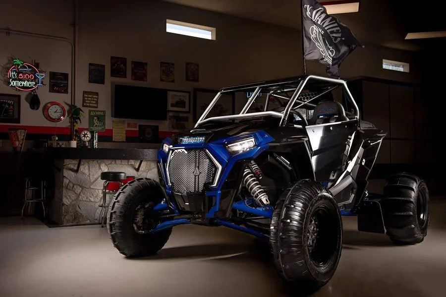 A rugged off-road vehicle with large tires, a roll cage, and a flag, parked indoors in a garage or showroom setting.