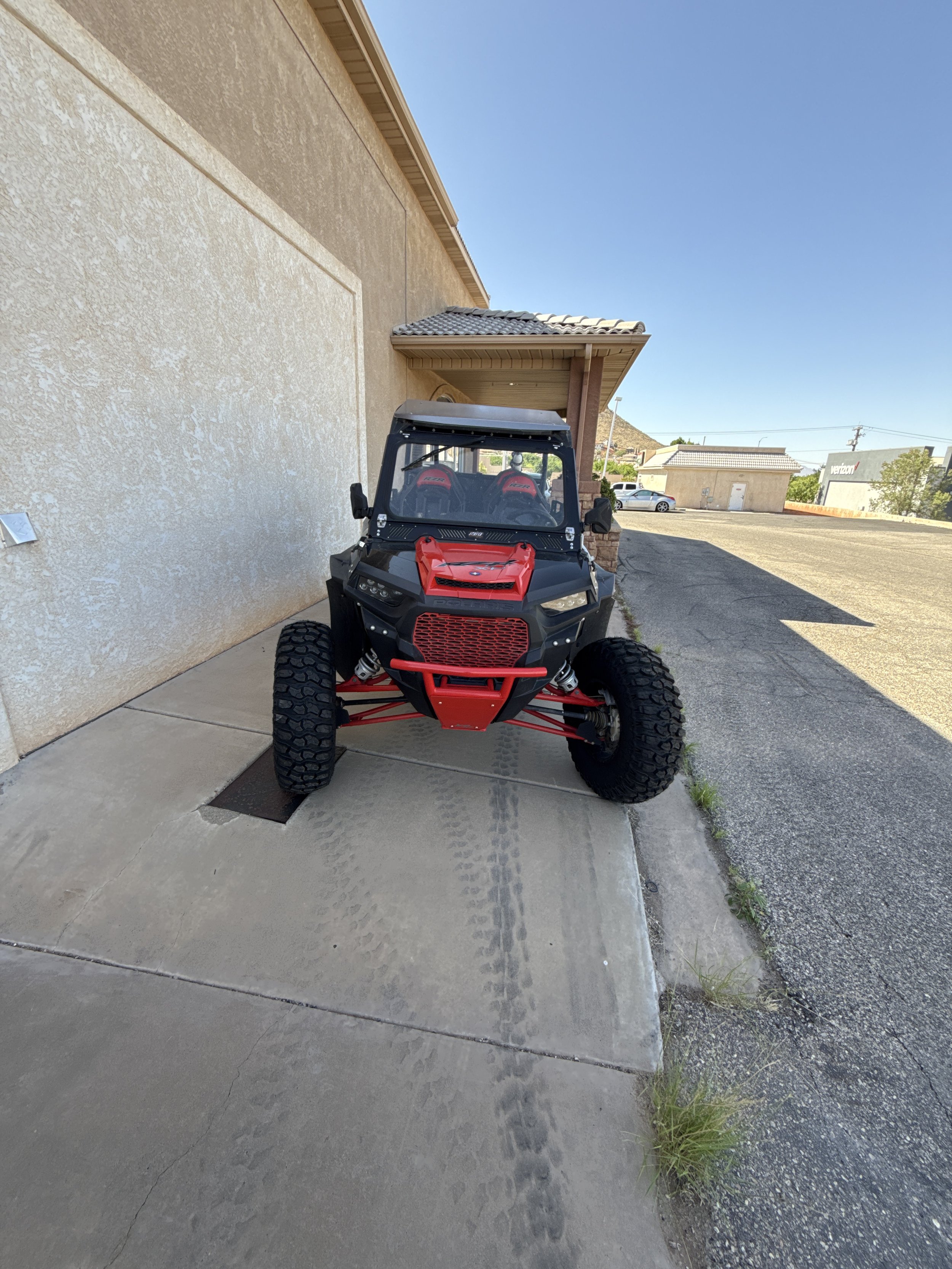 A black off-road dune buggy with large tires, exposed suspension, and a metal frame, parked on a concrete sidewalk in front of a beige wall with two flags on top.