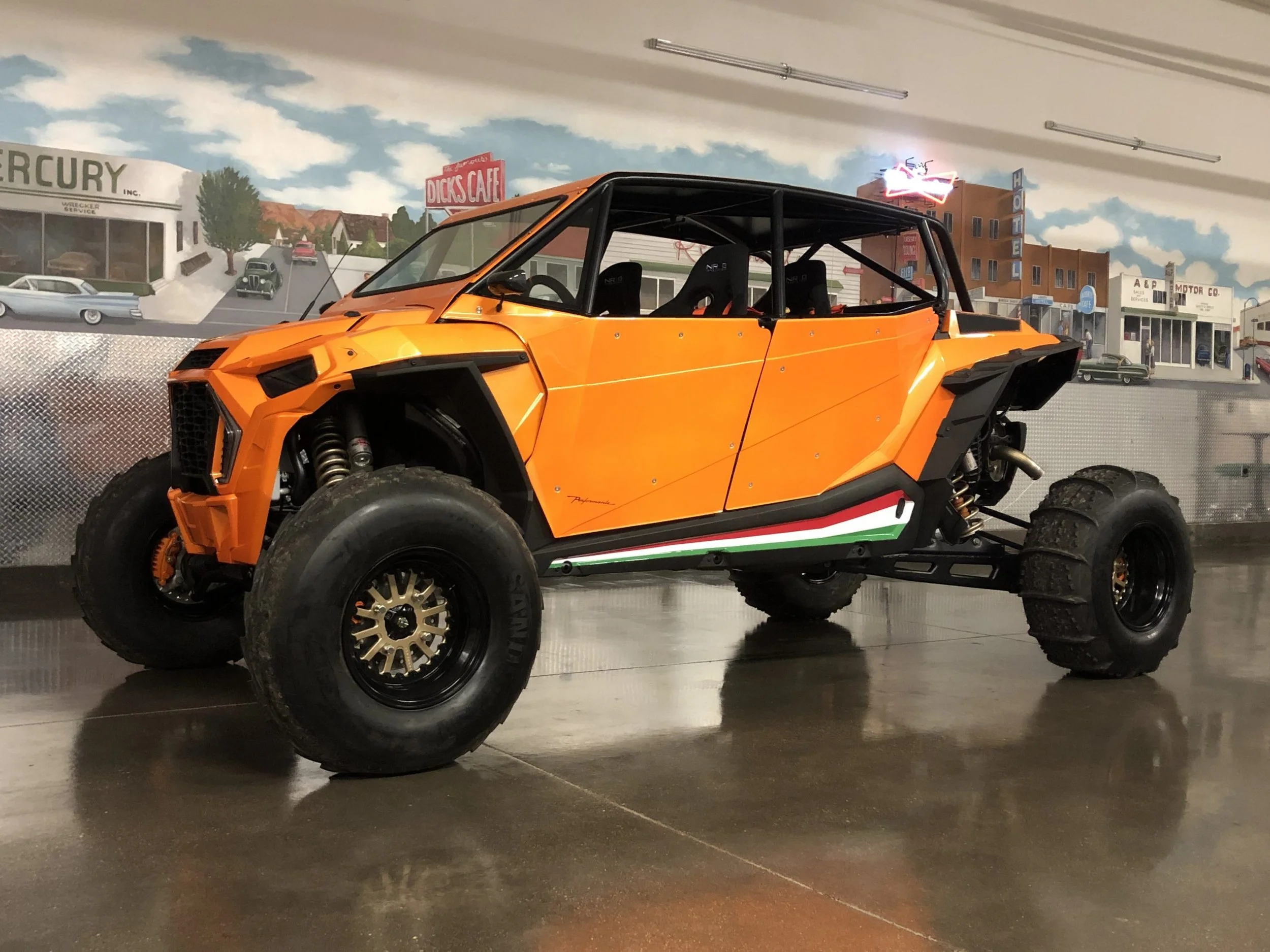 An off-road vehicle with large tires and an orange body parked indoors against a mural of a town scene.