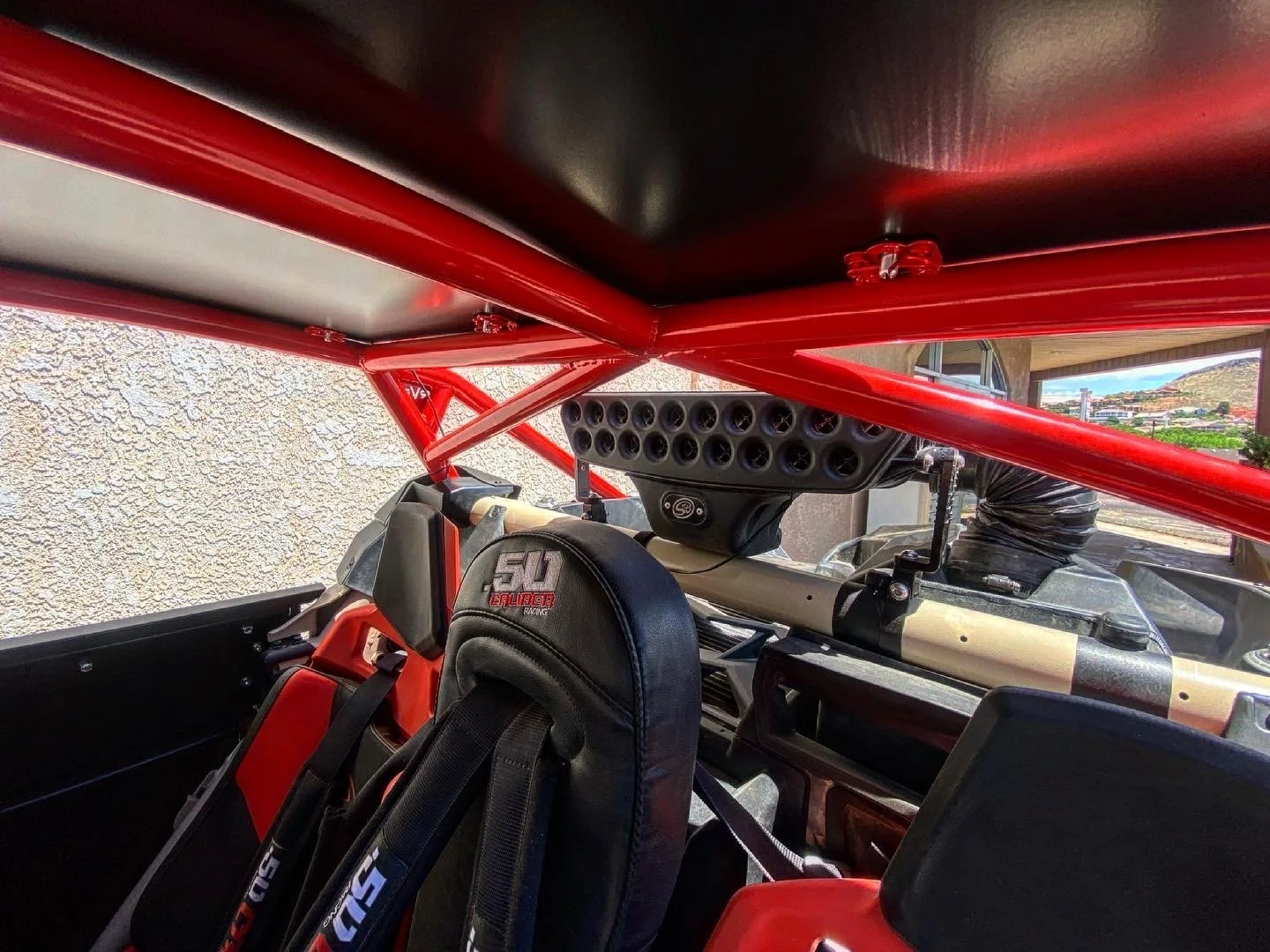 Interior of a race car, showing a racing seat with a harness and a roll cage, with an external landscape visible through the window.